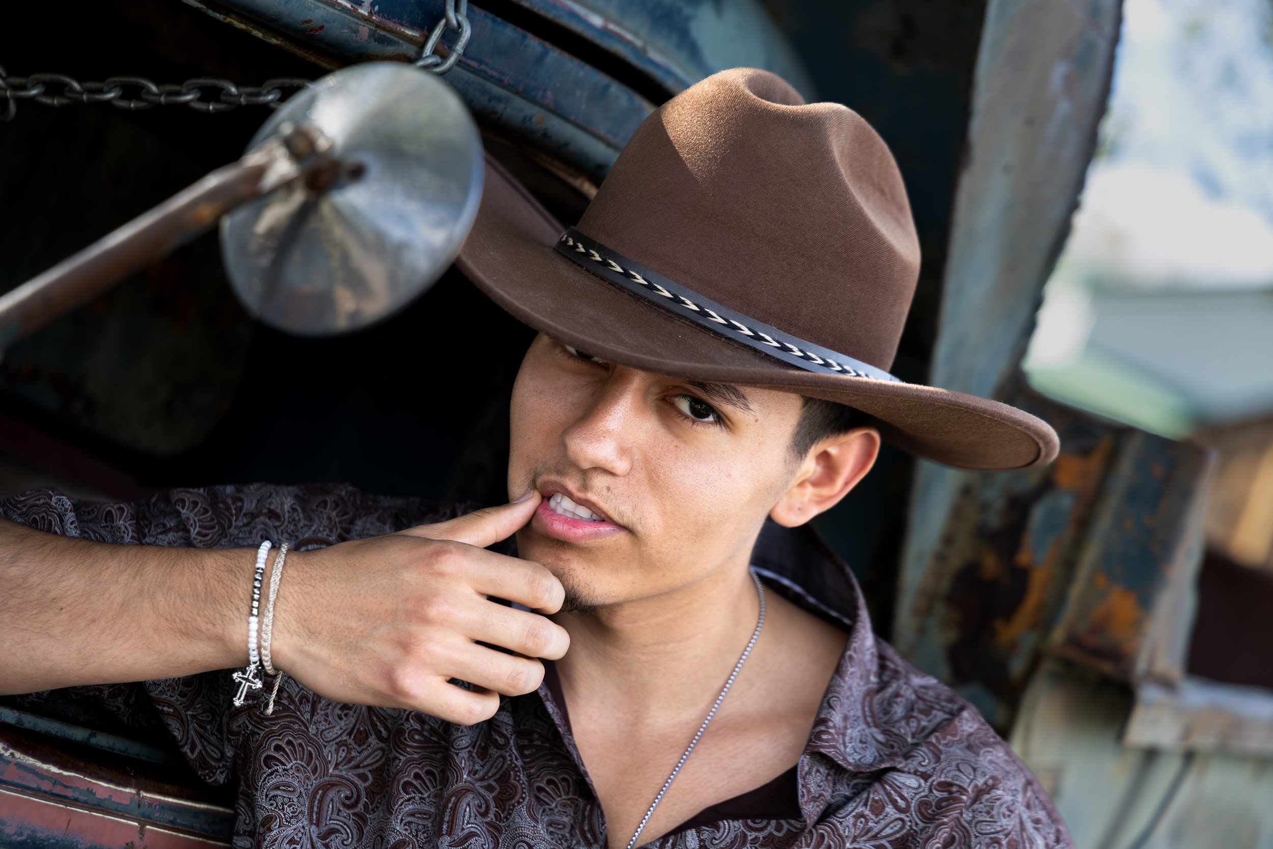 A man wearing a brown fedora hat with a black and white band, a patterned shirt, silver jewelry, and holding his finger to his lips, appears to be sitting under a rustic, weathered wooden structure.