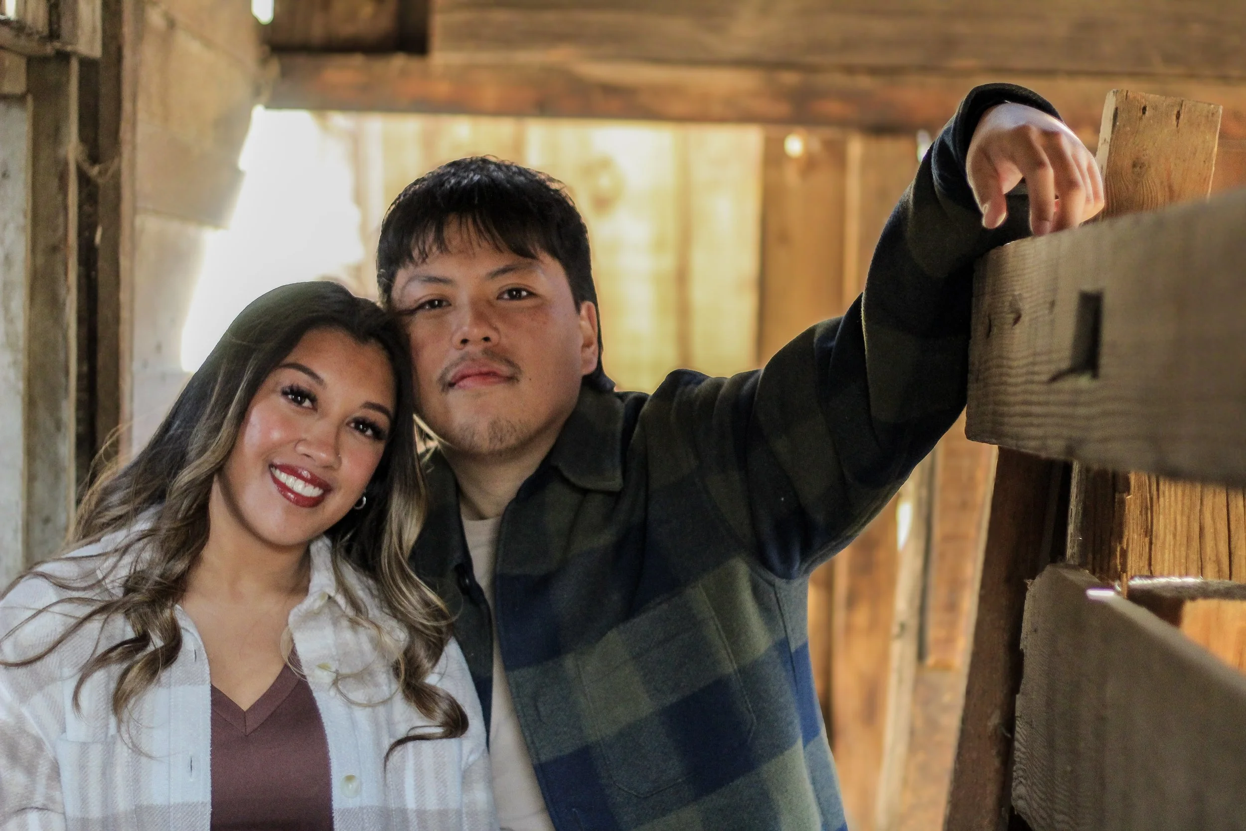 A smiling couple leaning close together inside a rustic wooden structure.