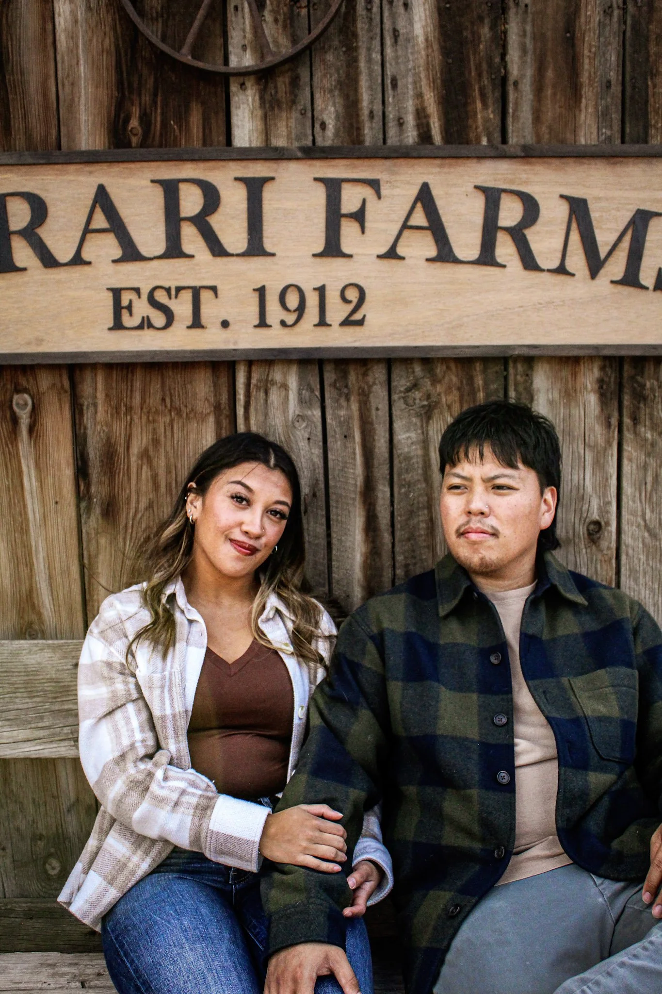 A woman and a man sitting in front of a wooden fence with a sign that reads 'FERRARI FARM EST. 1912'.