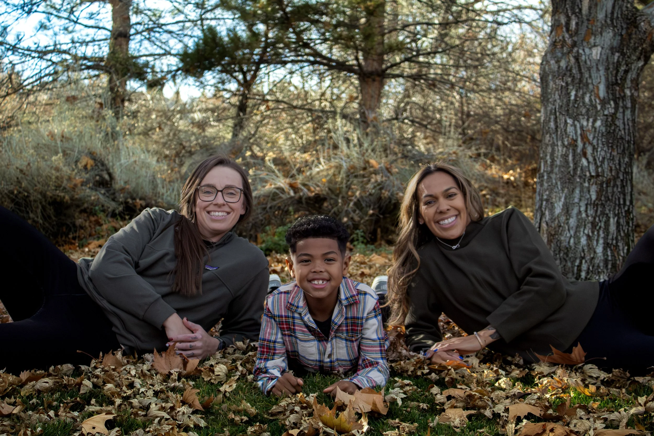 A smiling boy and two smiling women lying on the ground covered with fallen leaves in a wooded area during autumn.