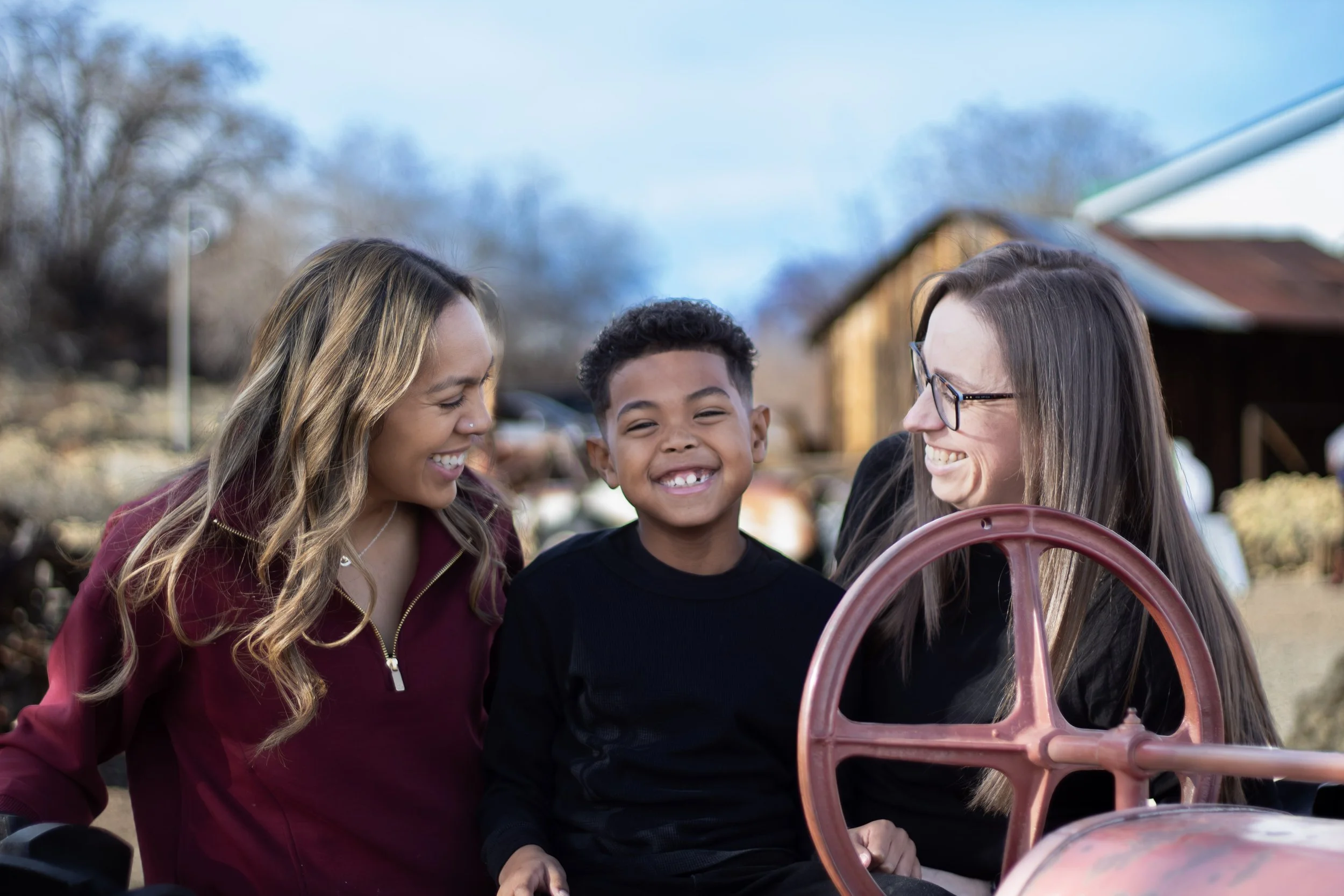 Three people smiling and laughing outdoors, with two women and one boy in the center, in front of farm equipment and a barn on a sunny day.