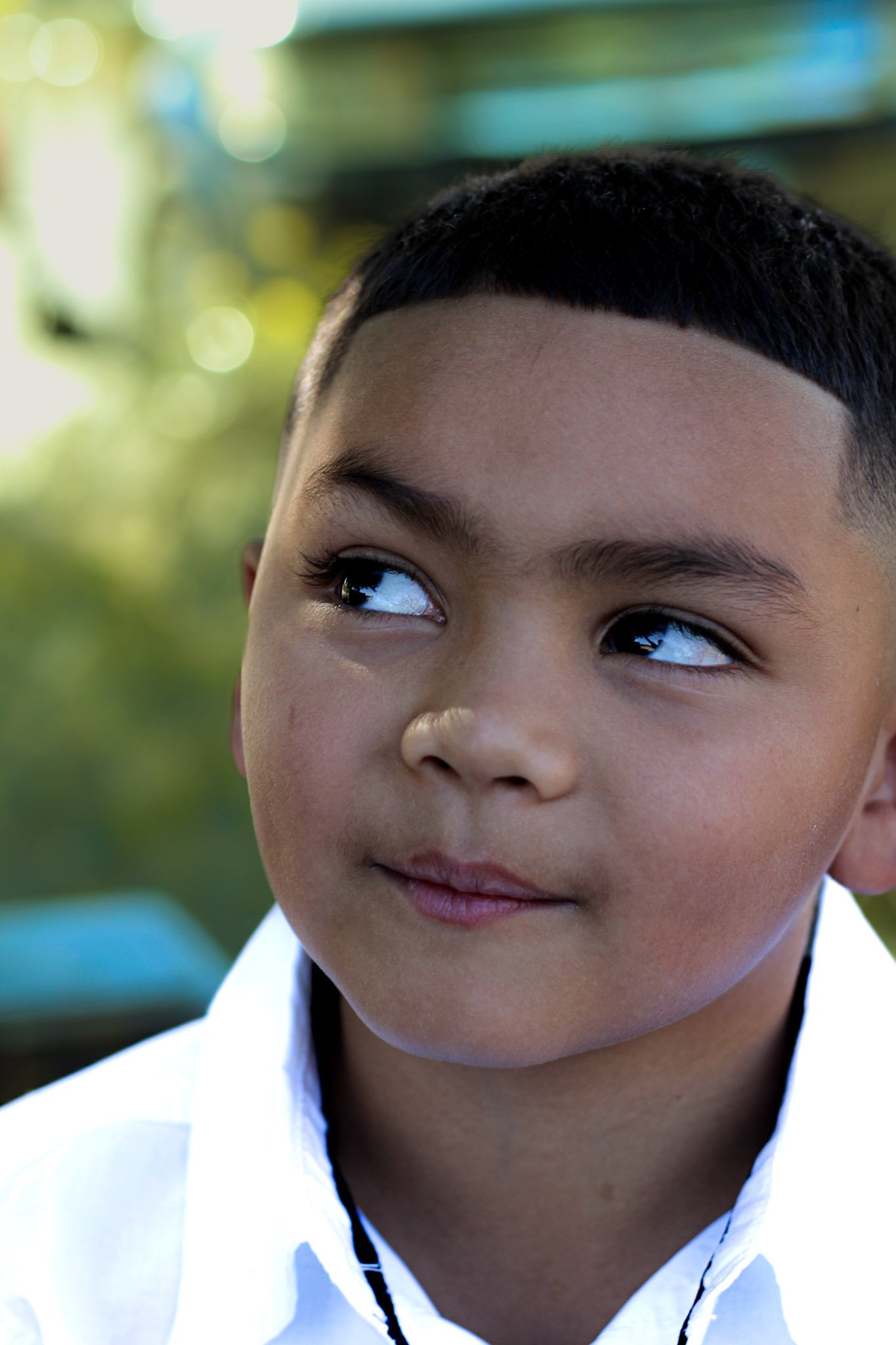 Close-up of a young boy with short dark hair, wearing a white shirt, looking to his right with a neutral expression, outdoor background with blurred greenery and sunlight.