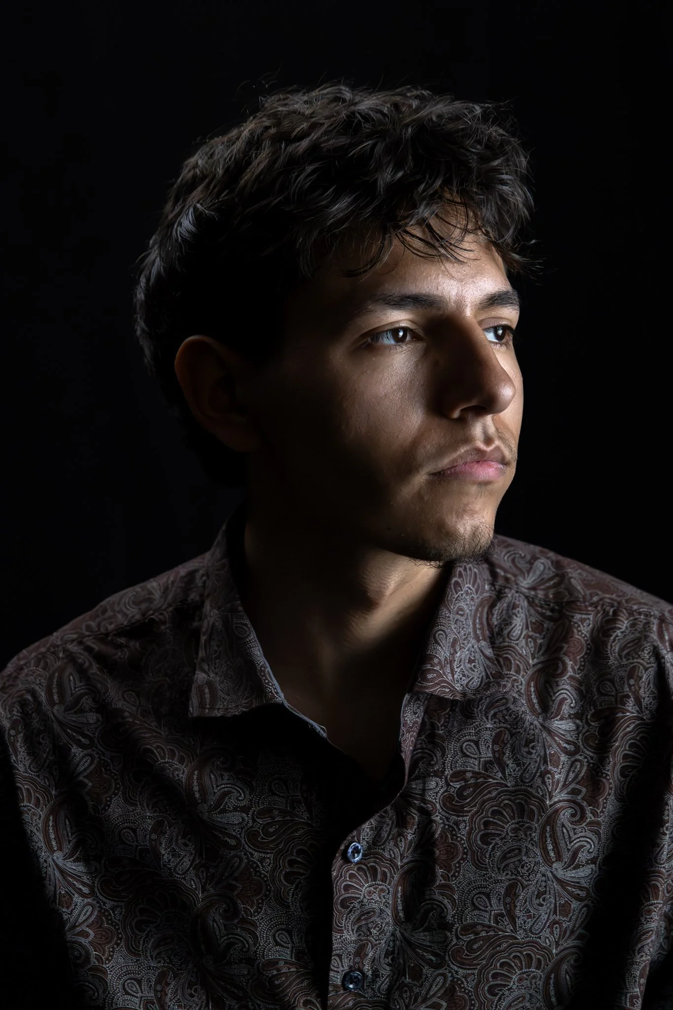 A young man with dark, curly hair and light skin gazing to the right against a black background, wearing a patterned button-up shirt.