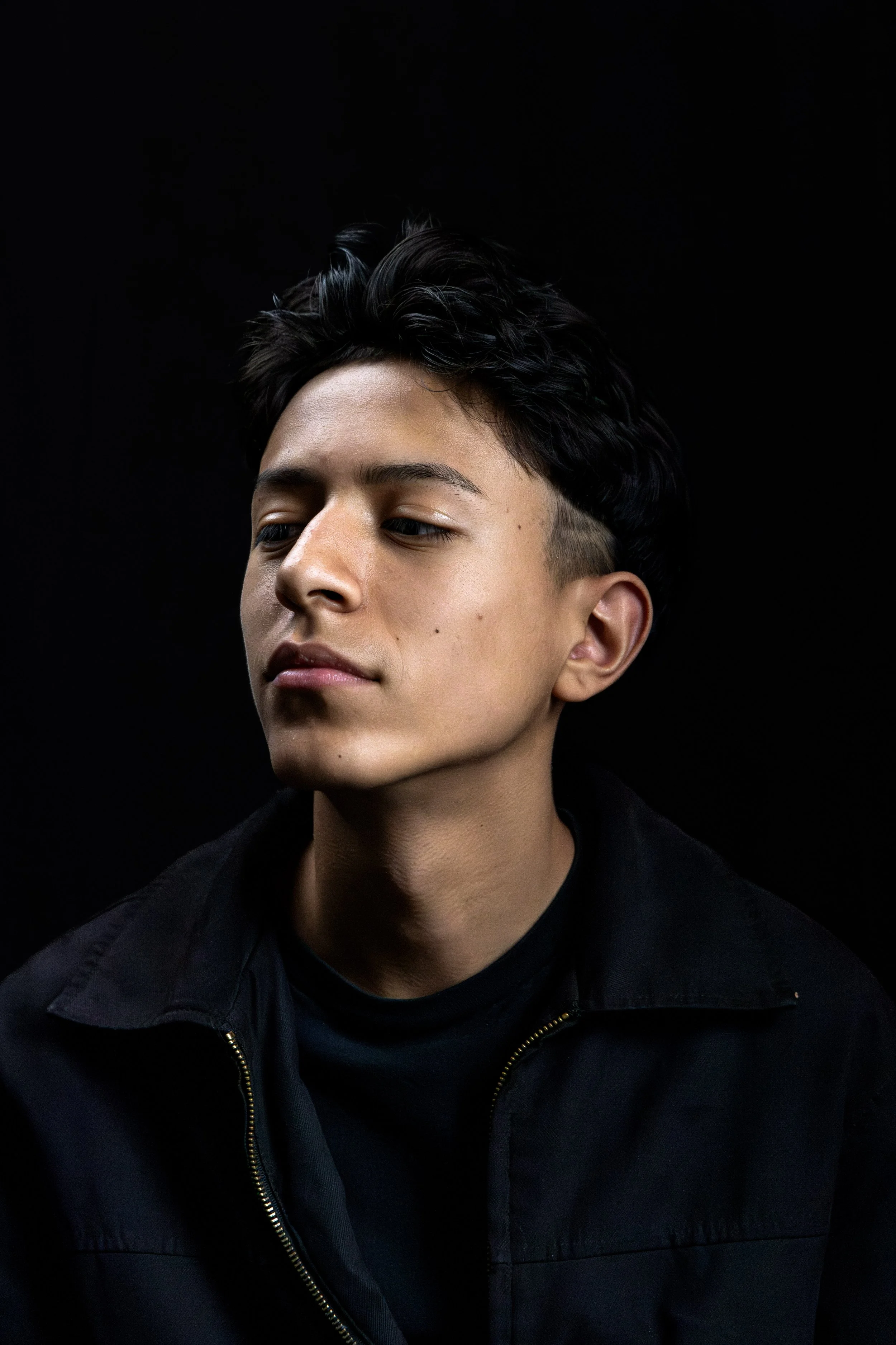 Portrait of a young man with dark hair and light skin, looking down and to the side, against a black background.