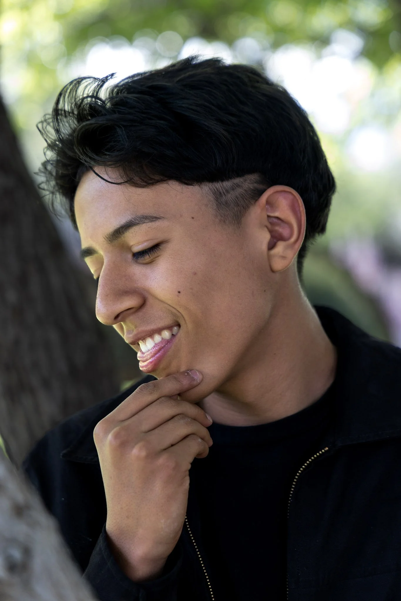 A young person with short black hair, smiling and touching their chin, outdoors with blurred green trees in the background.