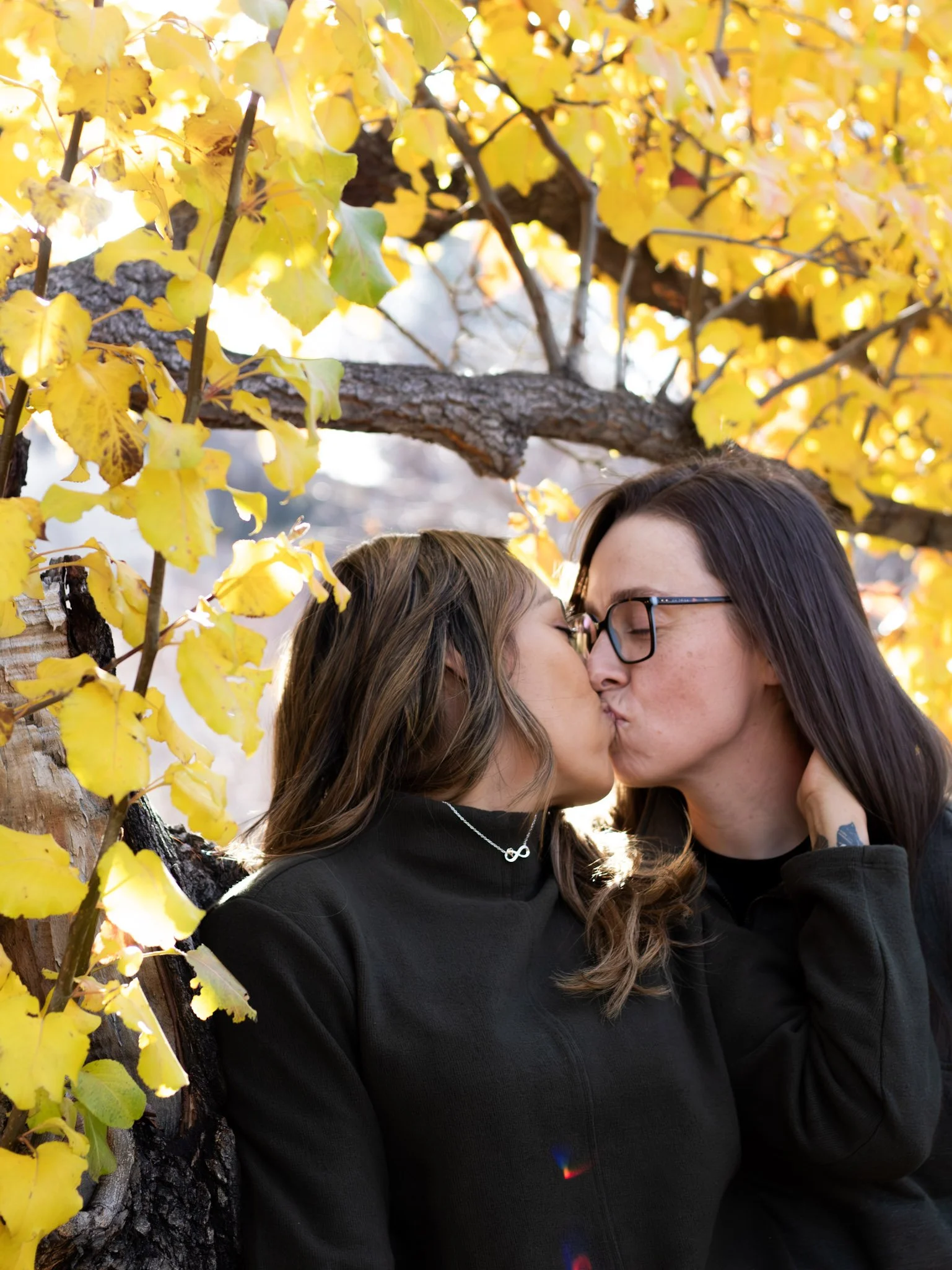 Two women kissing outdoors during fall, surrounded by yellow autumn leaves and tree branches.