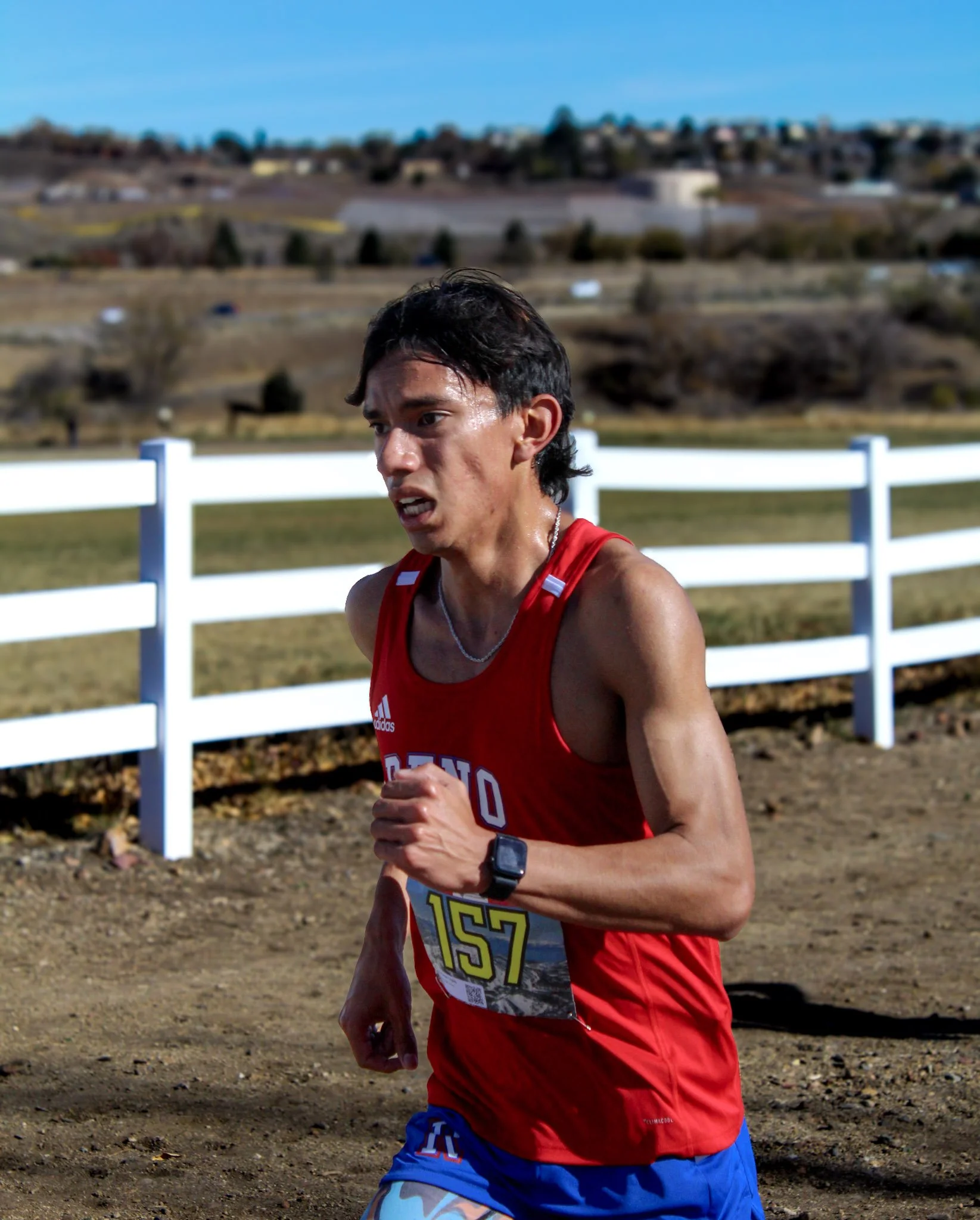 A male runner in a red sleeveless shirt and blue shorts running outdoors on a dirt trail, with a white fence and hilly landscape in the background.