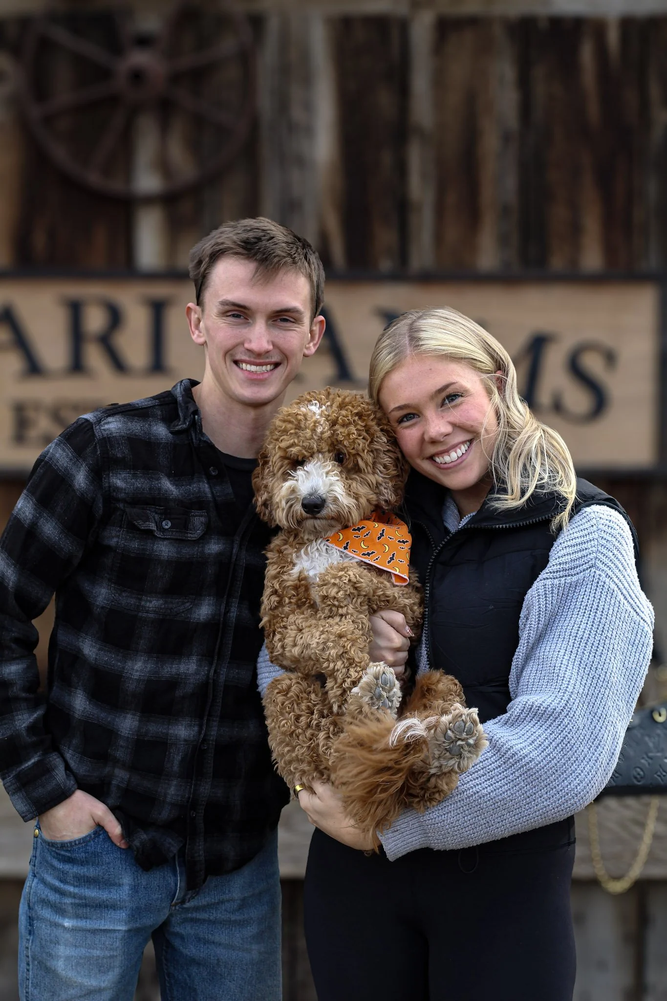 A smiling young couple standing outdoors in front of a rustic wooden background, holding a fluffy brown puppy with an orange bandana.