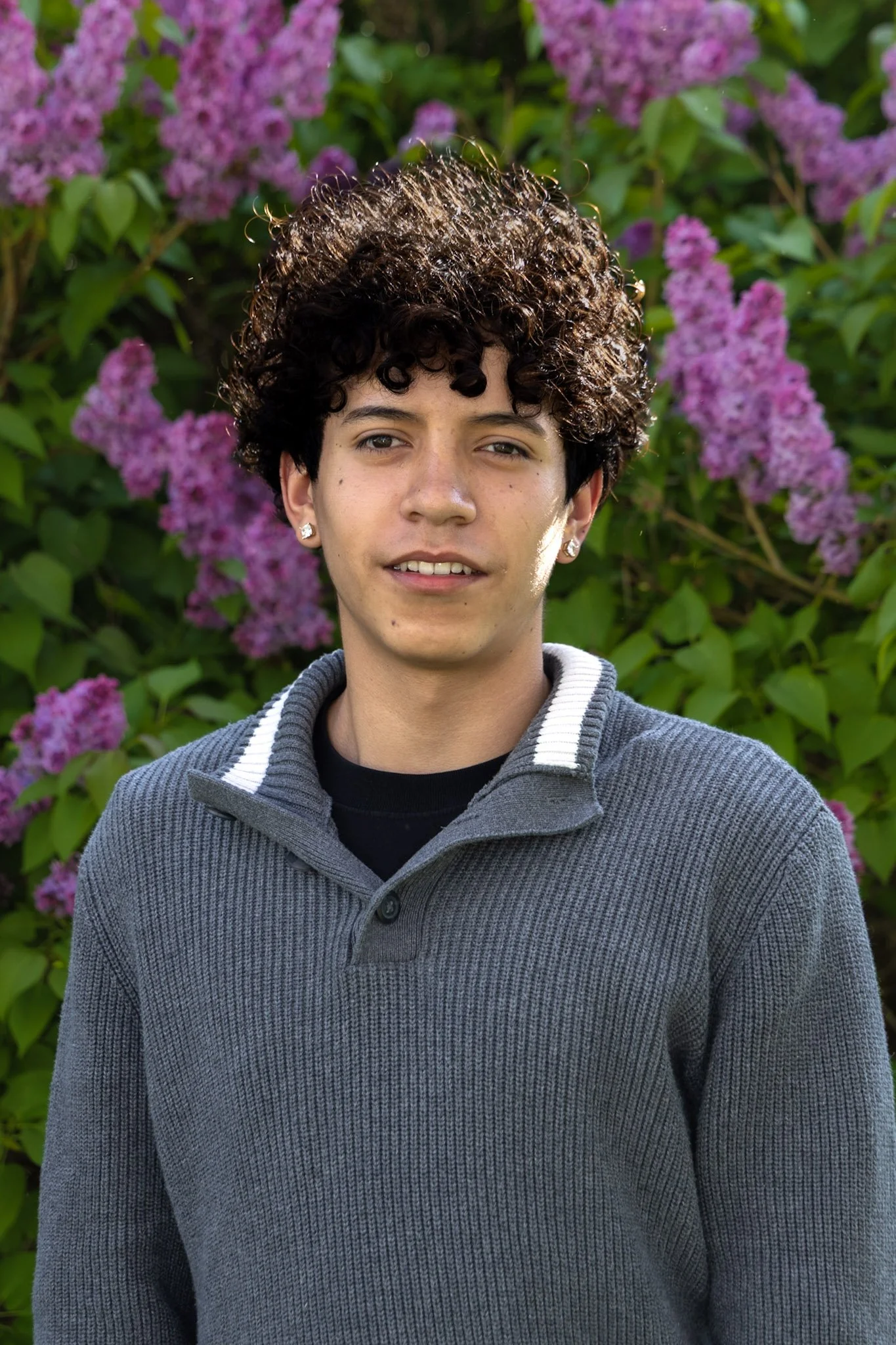 A young person with short, curly dark hair and earrings, wearing a grey sweater with a white stripe on the collar, standing outdoors in front of purple flowering bushes.