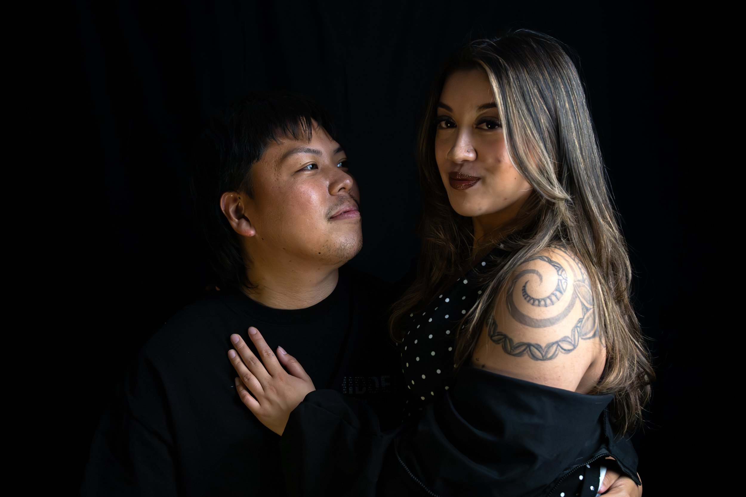 A man and a woman standing close together against a black background. The woman has long hair, dark lipstick, and tattoos on her shoulder. She is wearing a polka dot top and an open jacket, while the man wears a black shirt. They are gazing at each other with the woman smiling slightly.