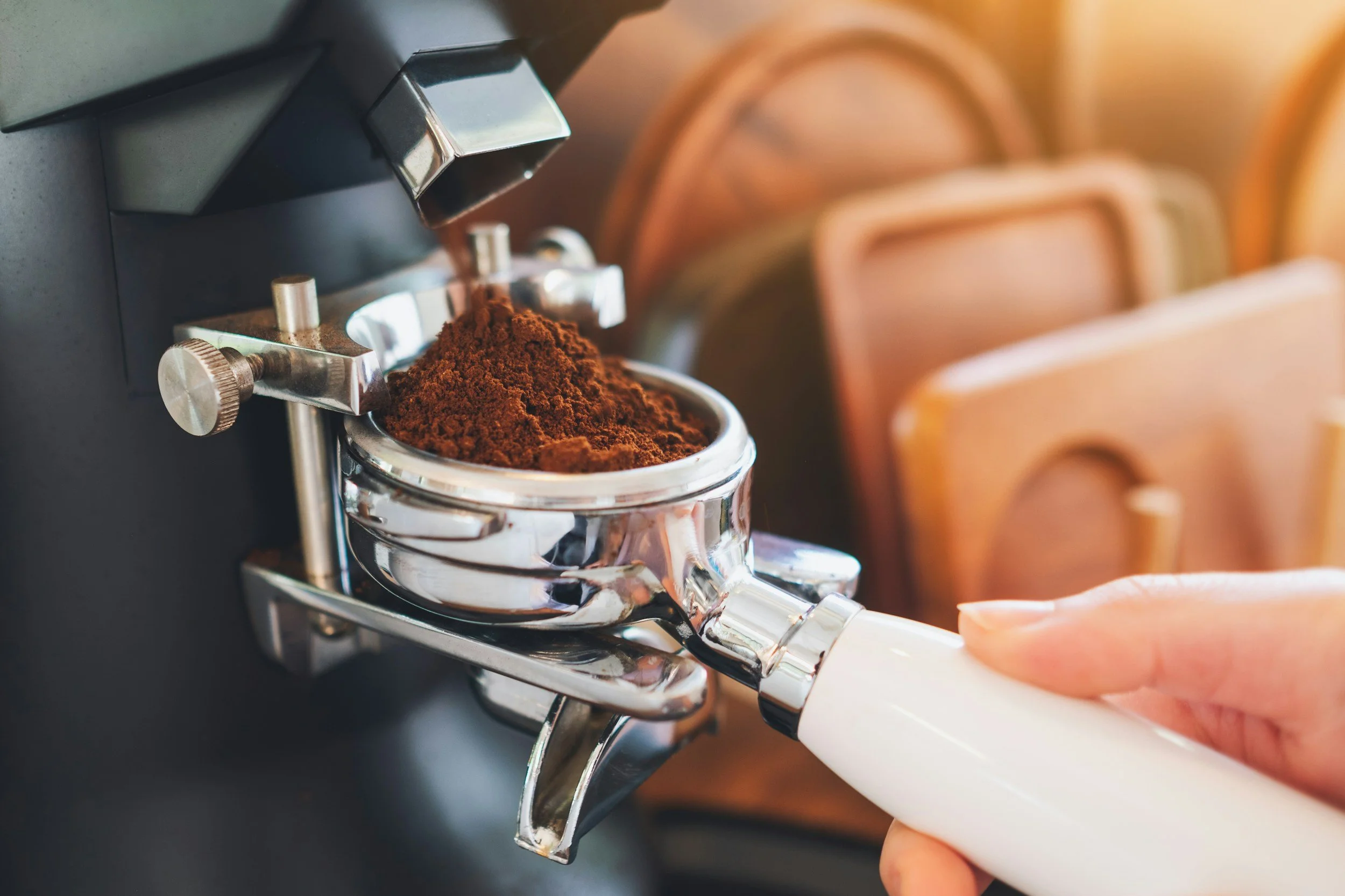A close-up of a coffee tamper pressing down ground coffee beans in a portafilter, with a hand holding the portafilter handle in a cafe setting.
