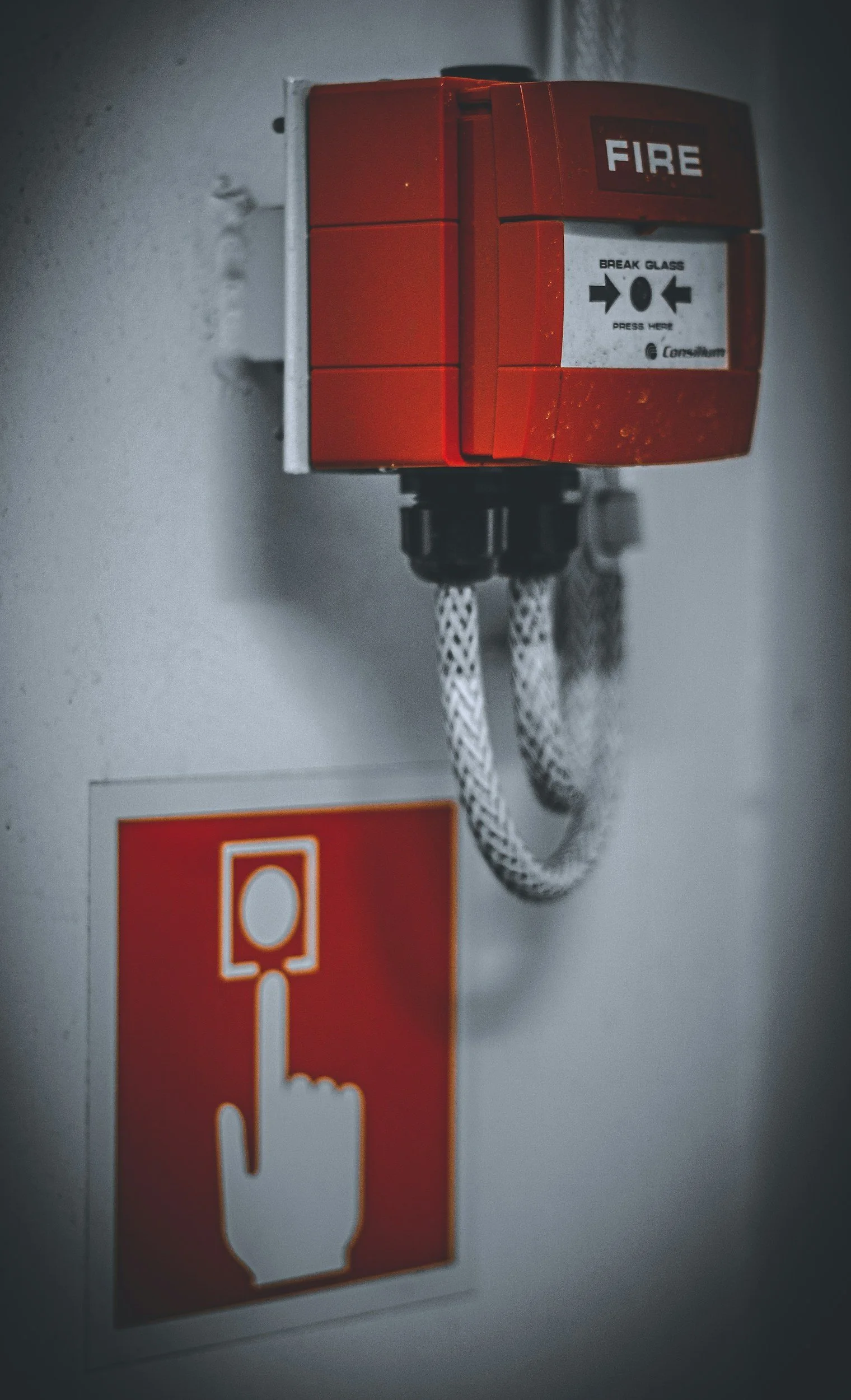 Red fire alarm pull station with a label indicating to break glass to press it, mounted on a gray wall. Below the alarm is a red sign with a white emergency icon.