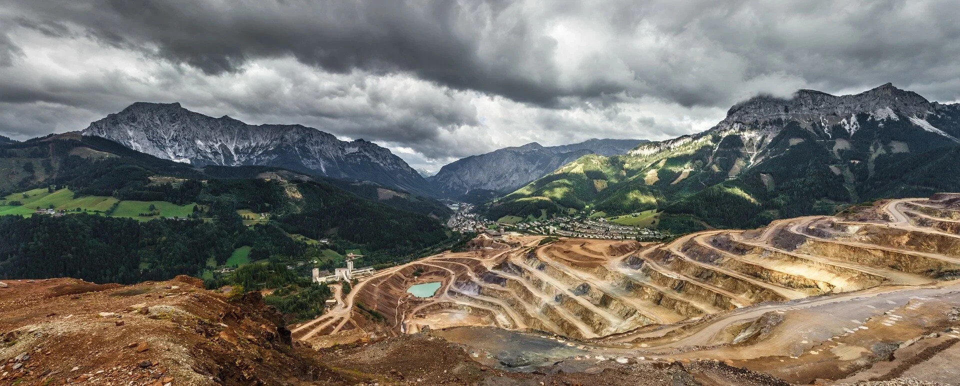A mountainous landscape with forested green hills and dark, rugged mountains under a cloudy sky. In the foreground, there is an open-pit mine with terraced levels and a small turquoise lake.