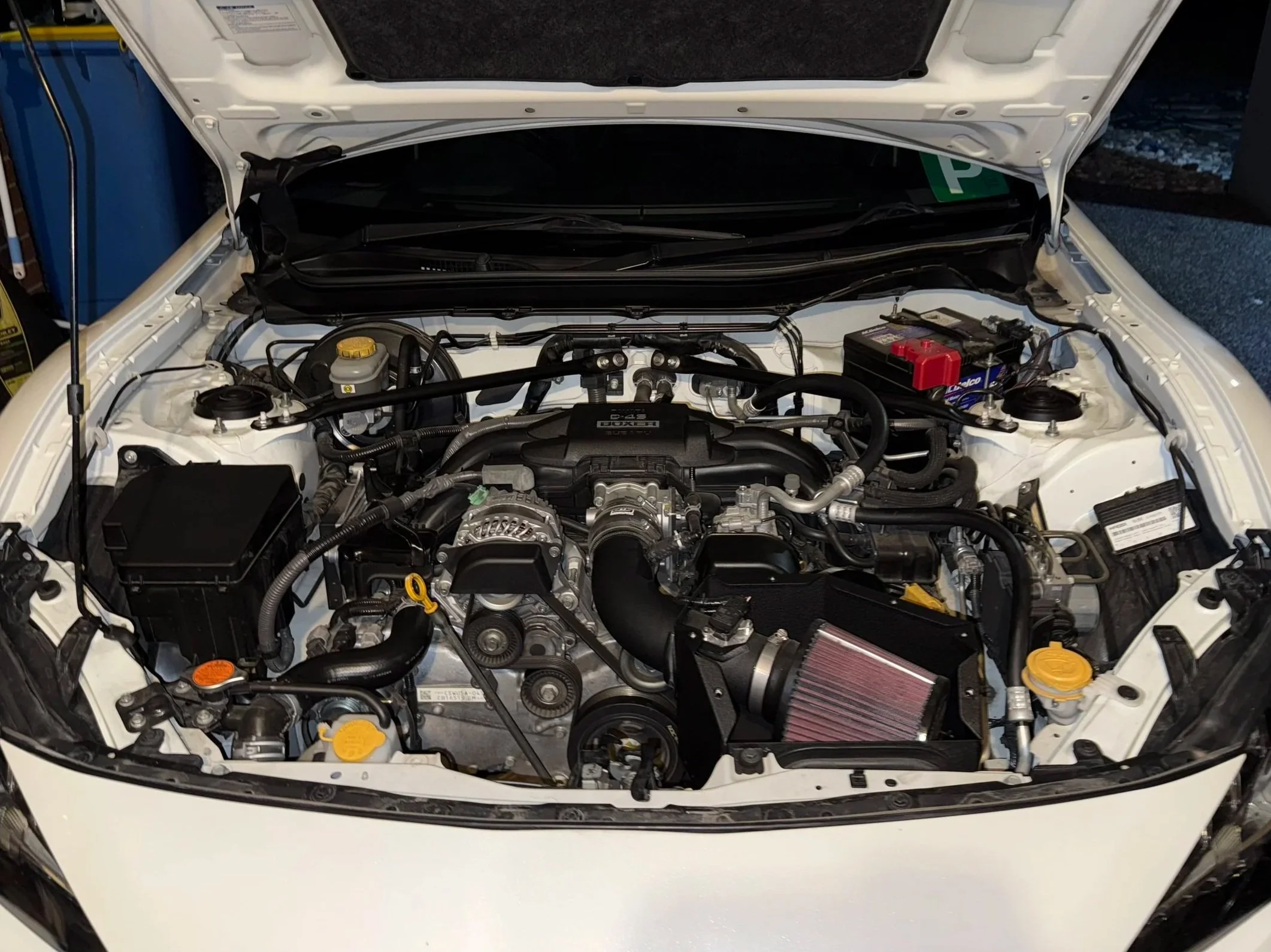 Inside view of a clean car engine bay showing various engine components, hoses, and a red-capped battery on the right side.
