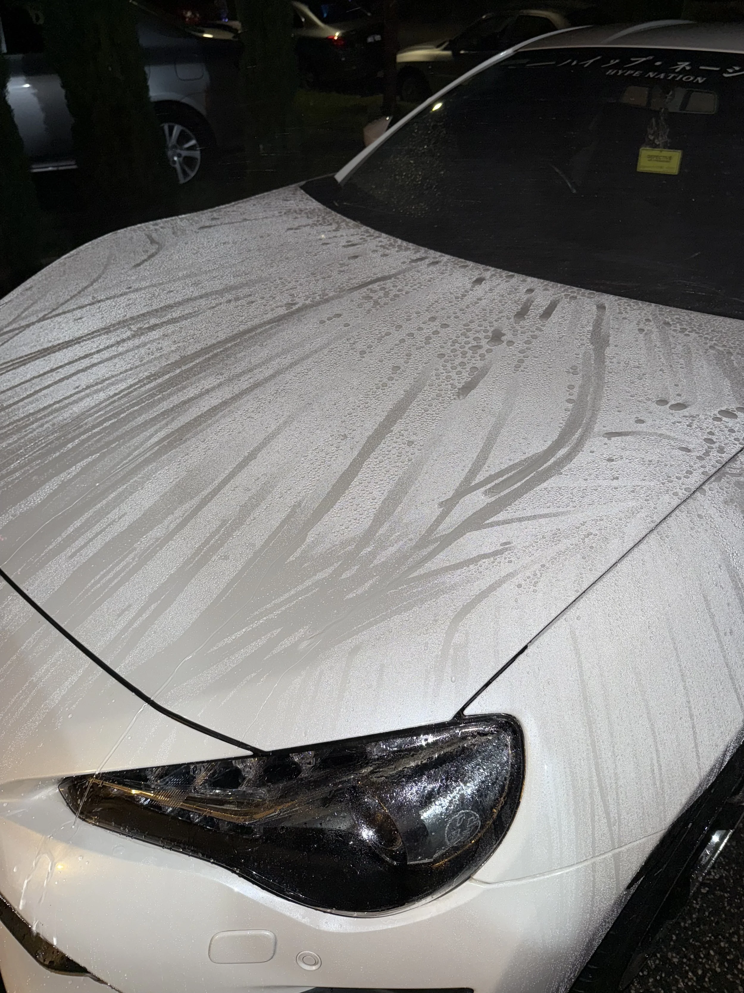 Rain-splattered white car hood with water streaks and droplets, parked in a lot at night.
