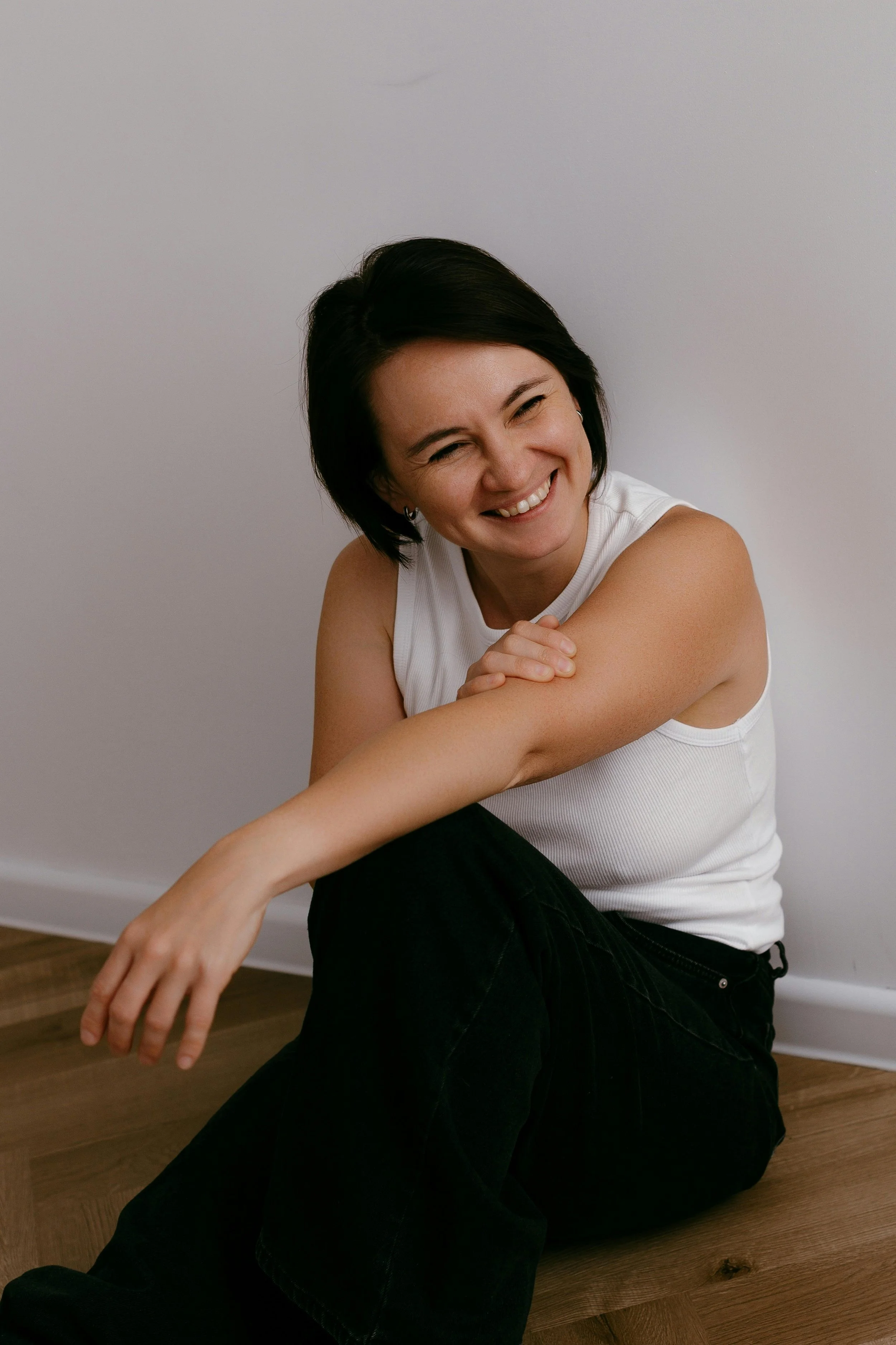 A woman with short black hair sitting on a wooden floor, smiling, wearing a sleeveless white top and black pants, against a plain white wall.