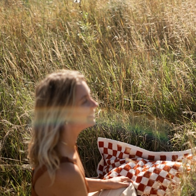 A woman with shoulder-length blonde hair sitting in a grassy field during sunset, with a checkered blanket beside her.