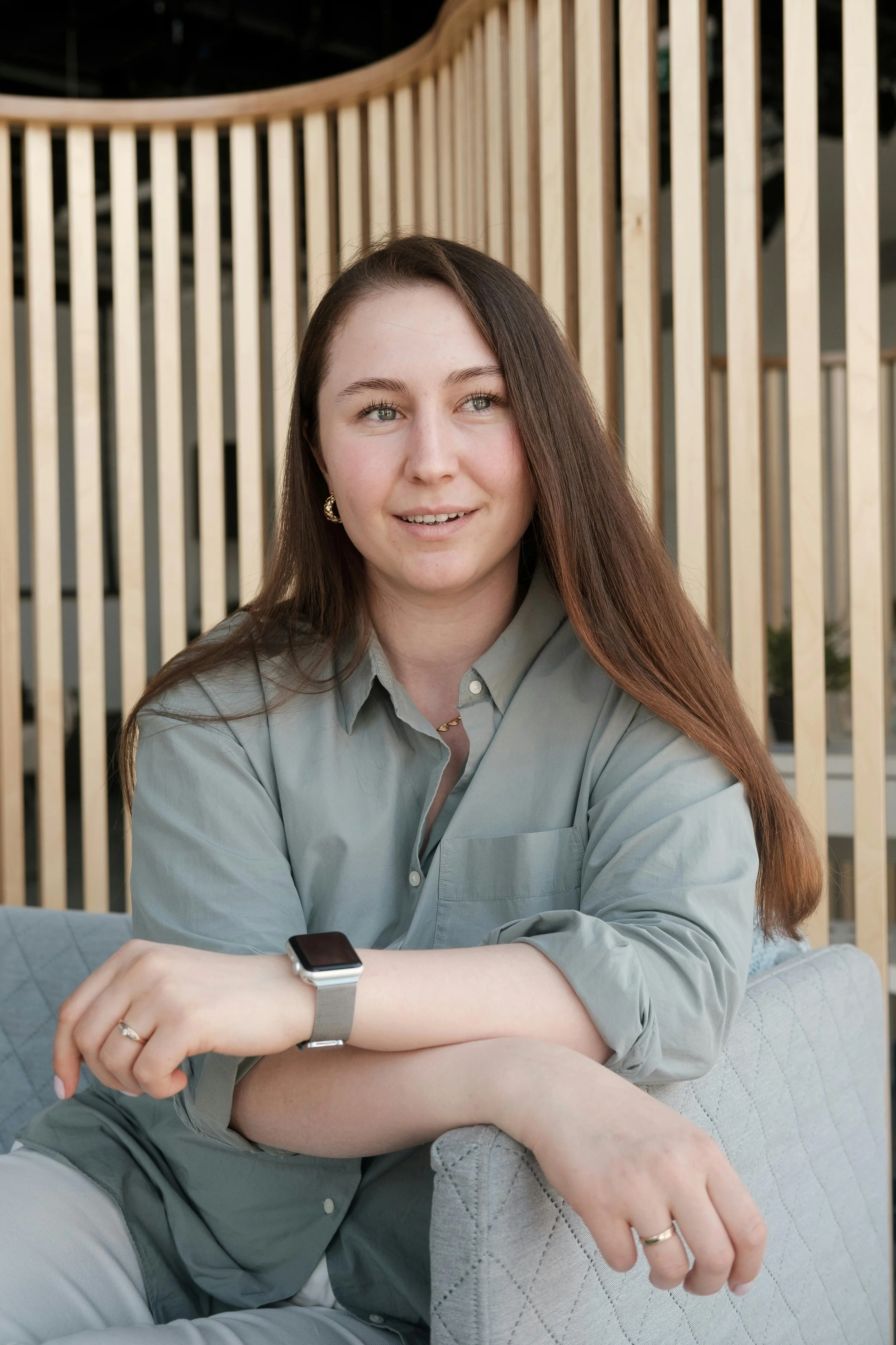 Close-up of a woman with long brown hair, wearing a gray button-up shirt, sitting on a light-colored quilted chair with her arms crossed, smiling slightly, in front of a wooden slat divider.