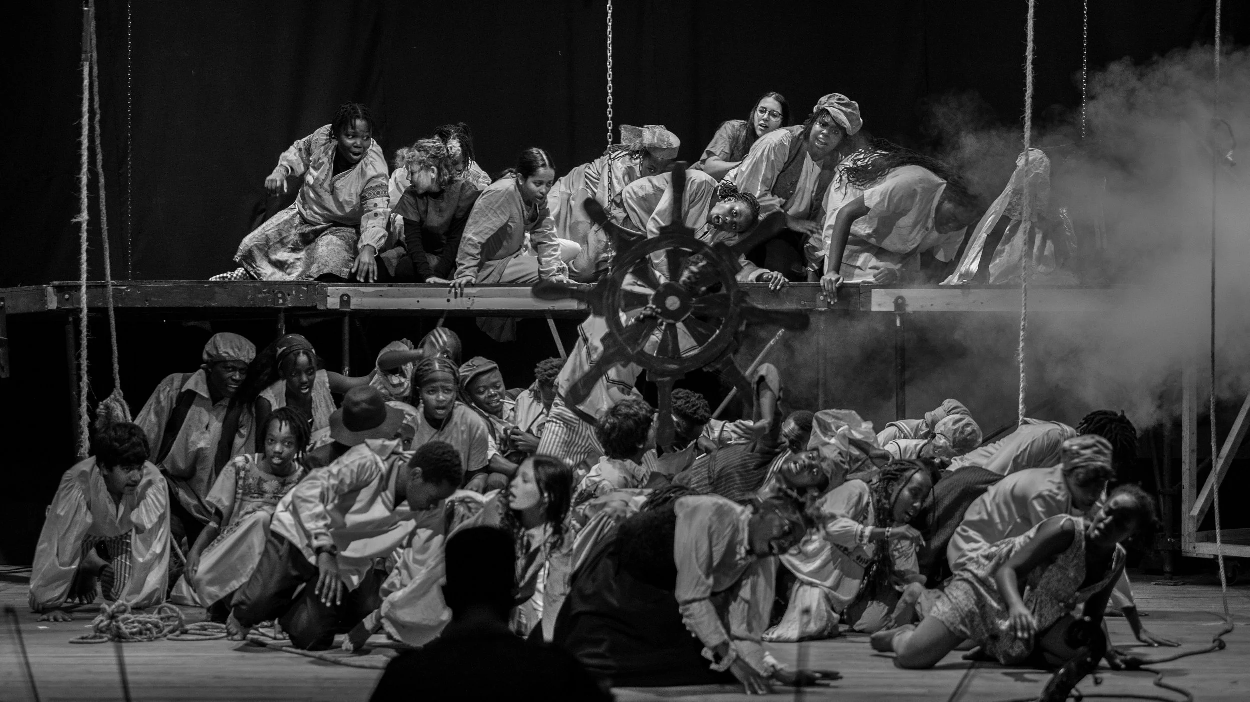 A black and white photo of a theatrical scene with numerous actors on stage, some on the upper level and others on the lower level, wearing costumes. Smoke effects and a large wheel prop are visible, creating a dramatic atmosphere.