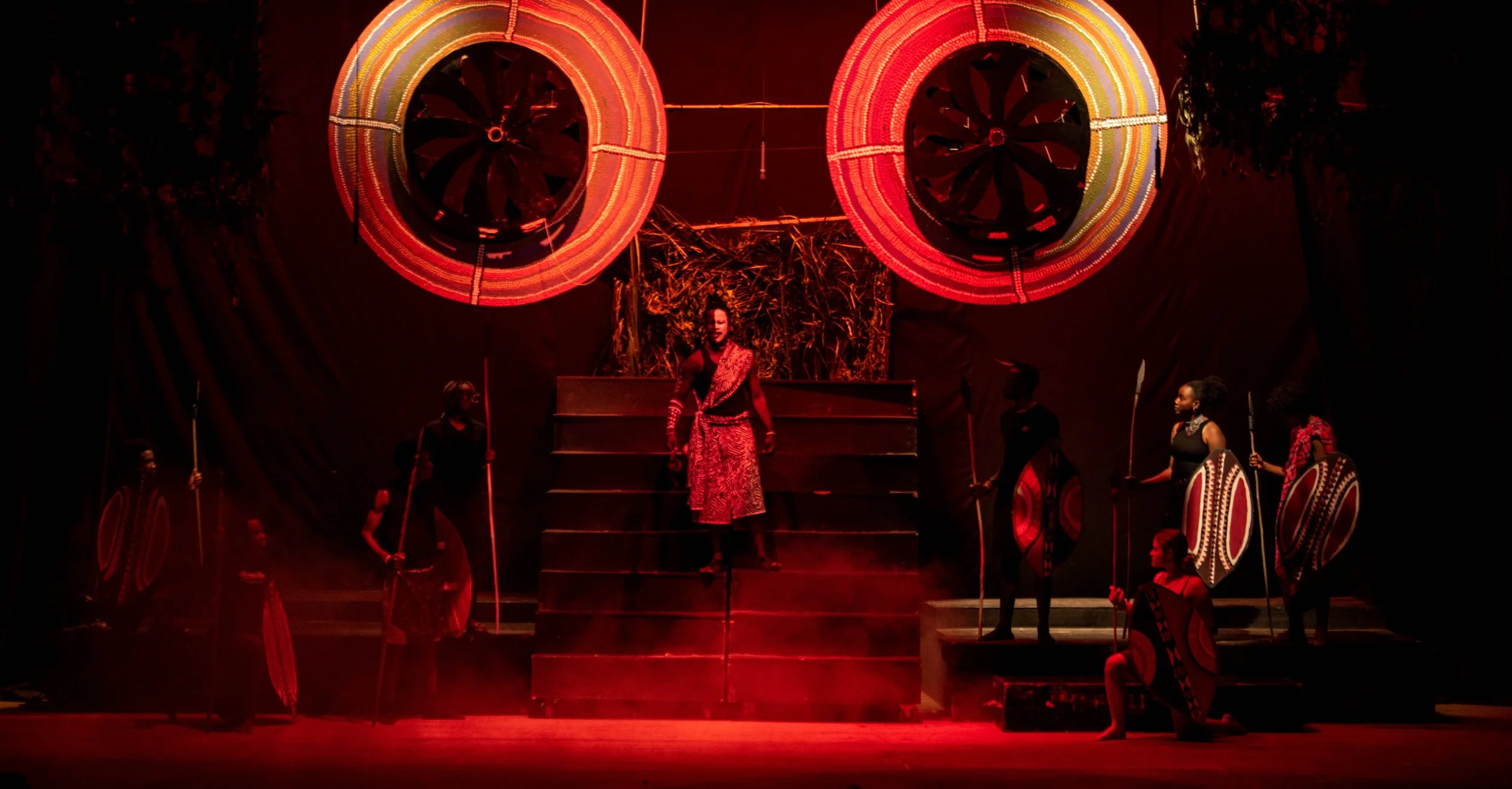 Performers in traditional African attire on stage, with two large circular woven decorations hanging above, illuminated in red.