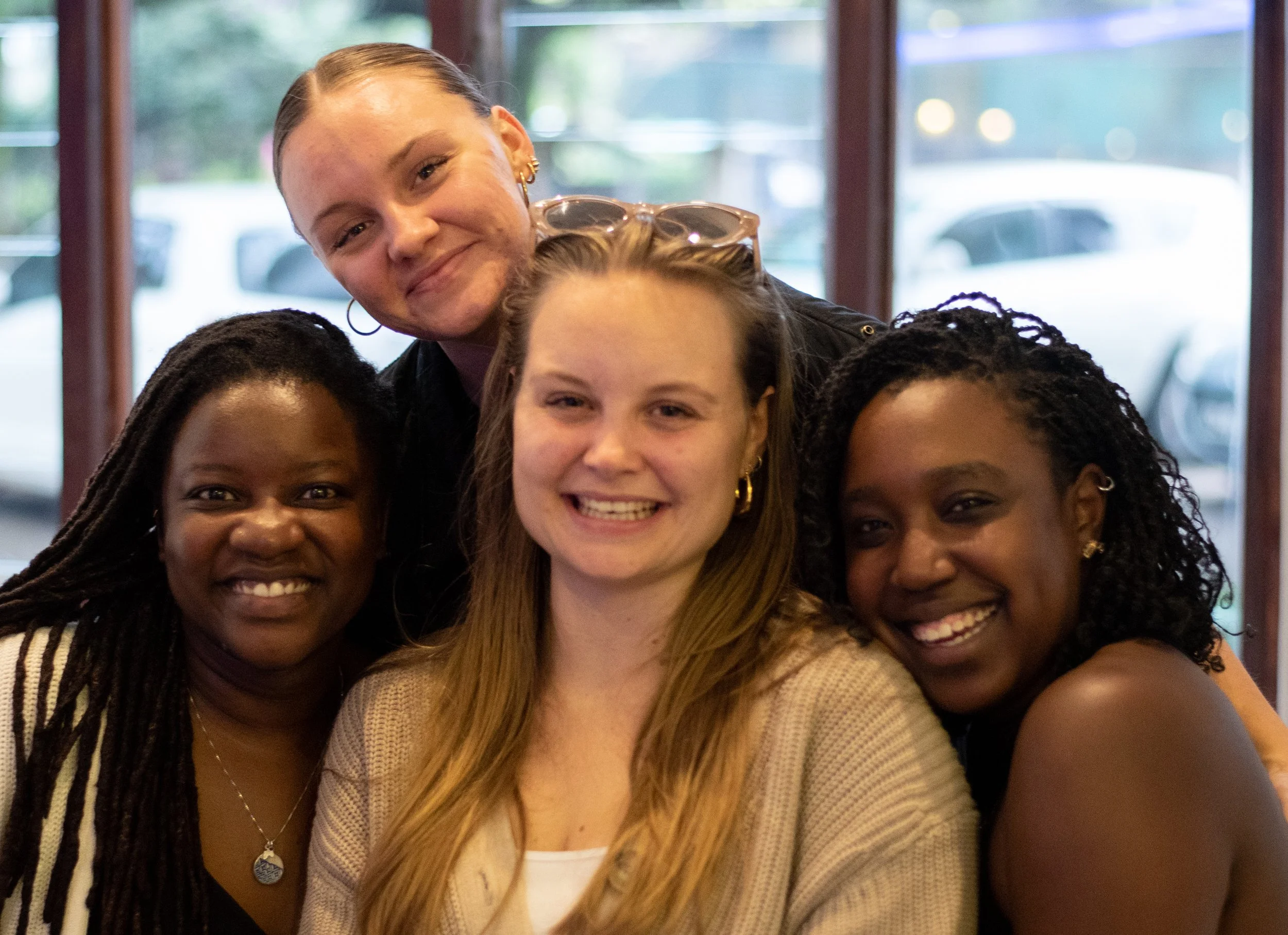 Group of five women smiling together indoors with large windows in the background.