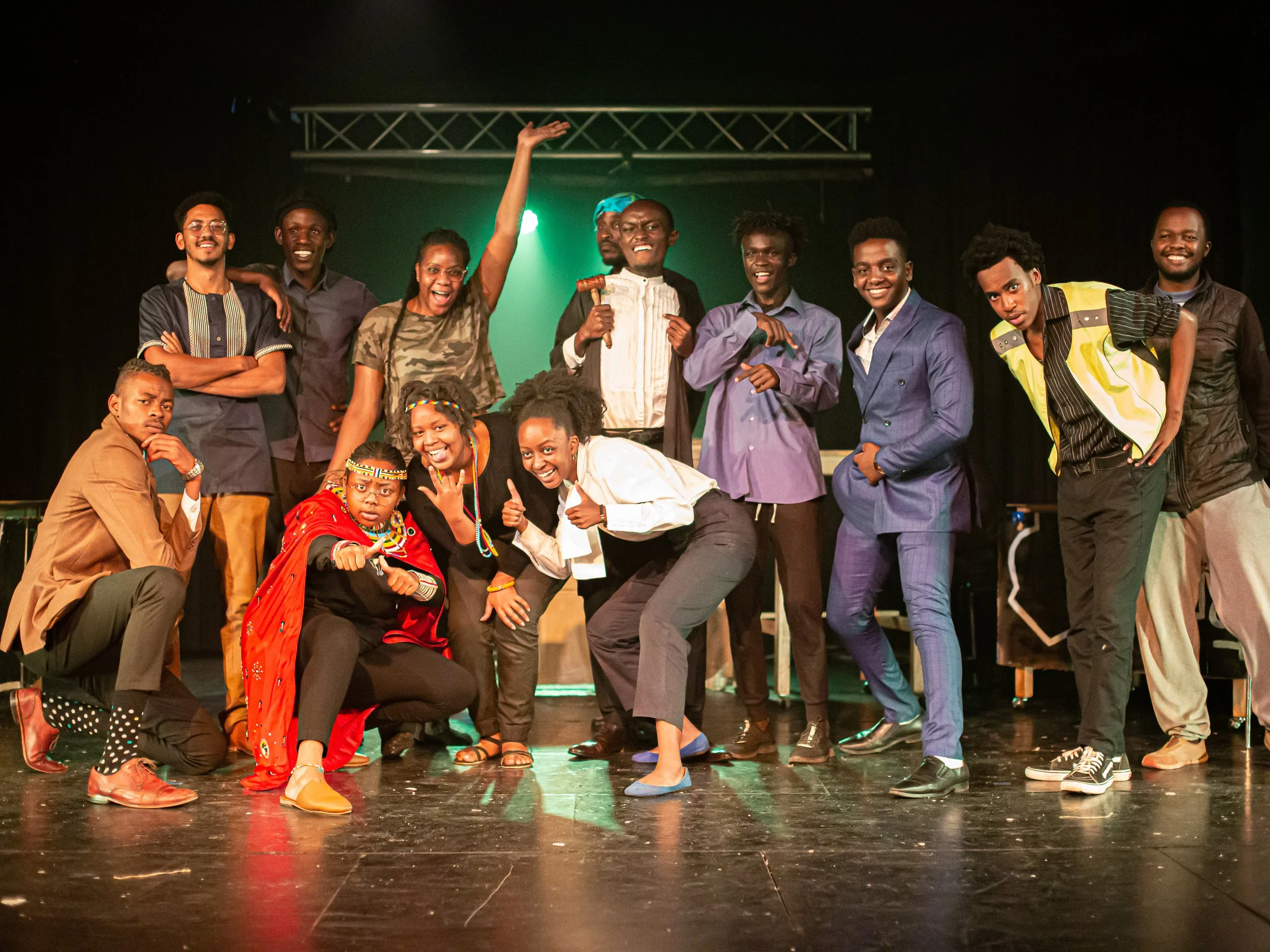 Group of diverse people posing on a stage, some wearing traditional and casual clothing, with a lighting setup overhead.