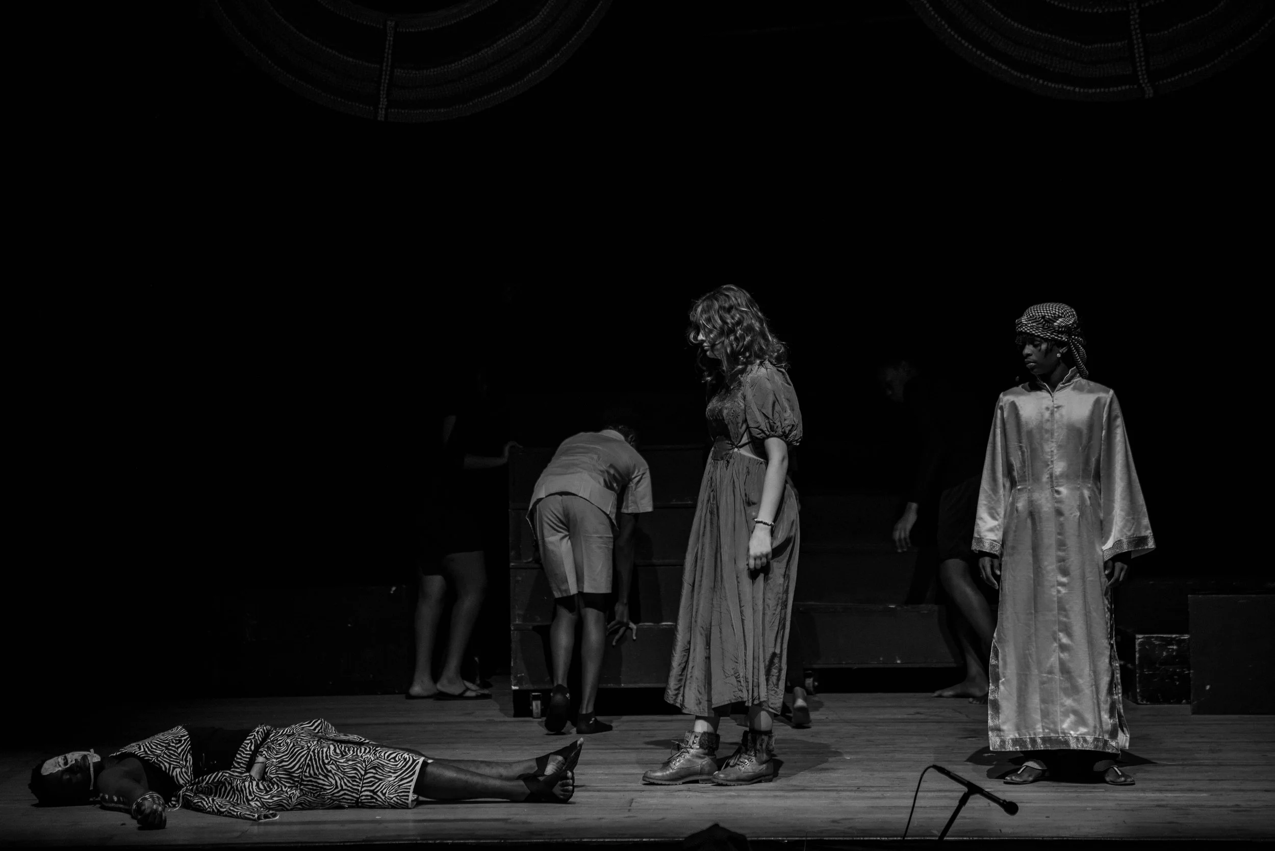 Black and white photo of a stage scene with five people. One person is lying on the floor, and four others are standing or bent over. The scene appears to be a theatrical performance.