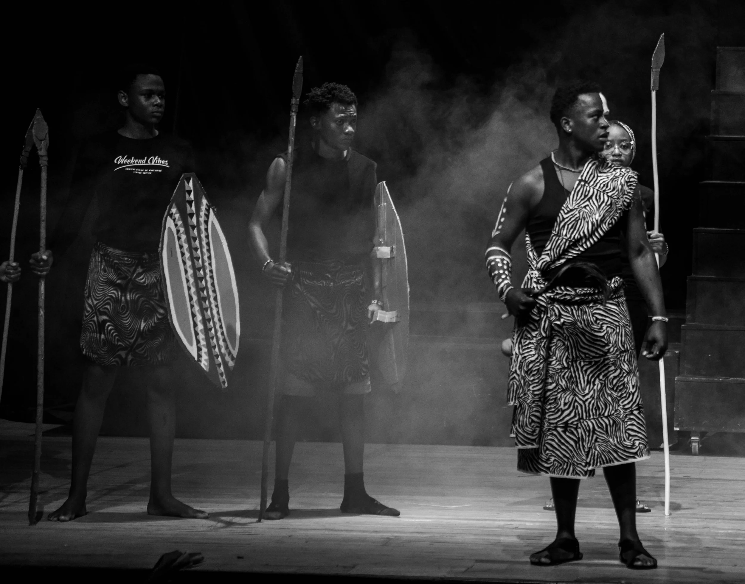 Four performers dressed in traditional African attire with face paint and carrying spears and shields, performing on stage with a smoky background.