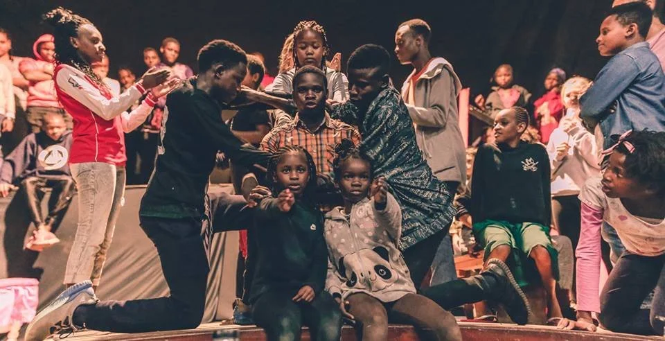 Group of children and adults gathered on a stage, some kneeling and some standing, with a dark background and stage lighting.