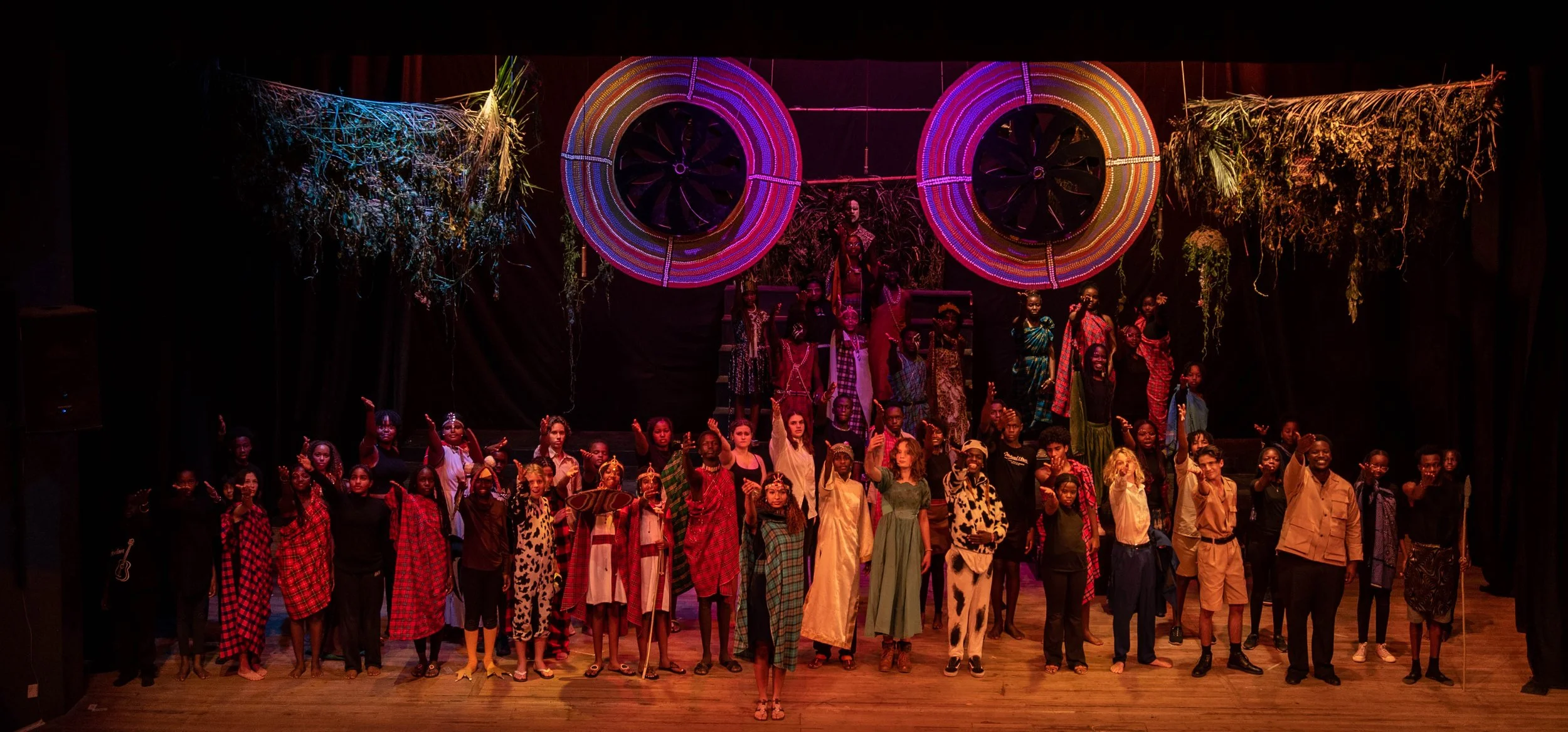 Group of performers on stage dressed in colorful costumes, some in traditional attire, with two large circular woven decorations, and greenery hanging from the ceiling.