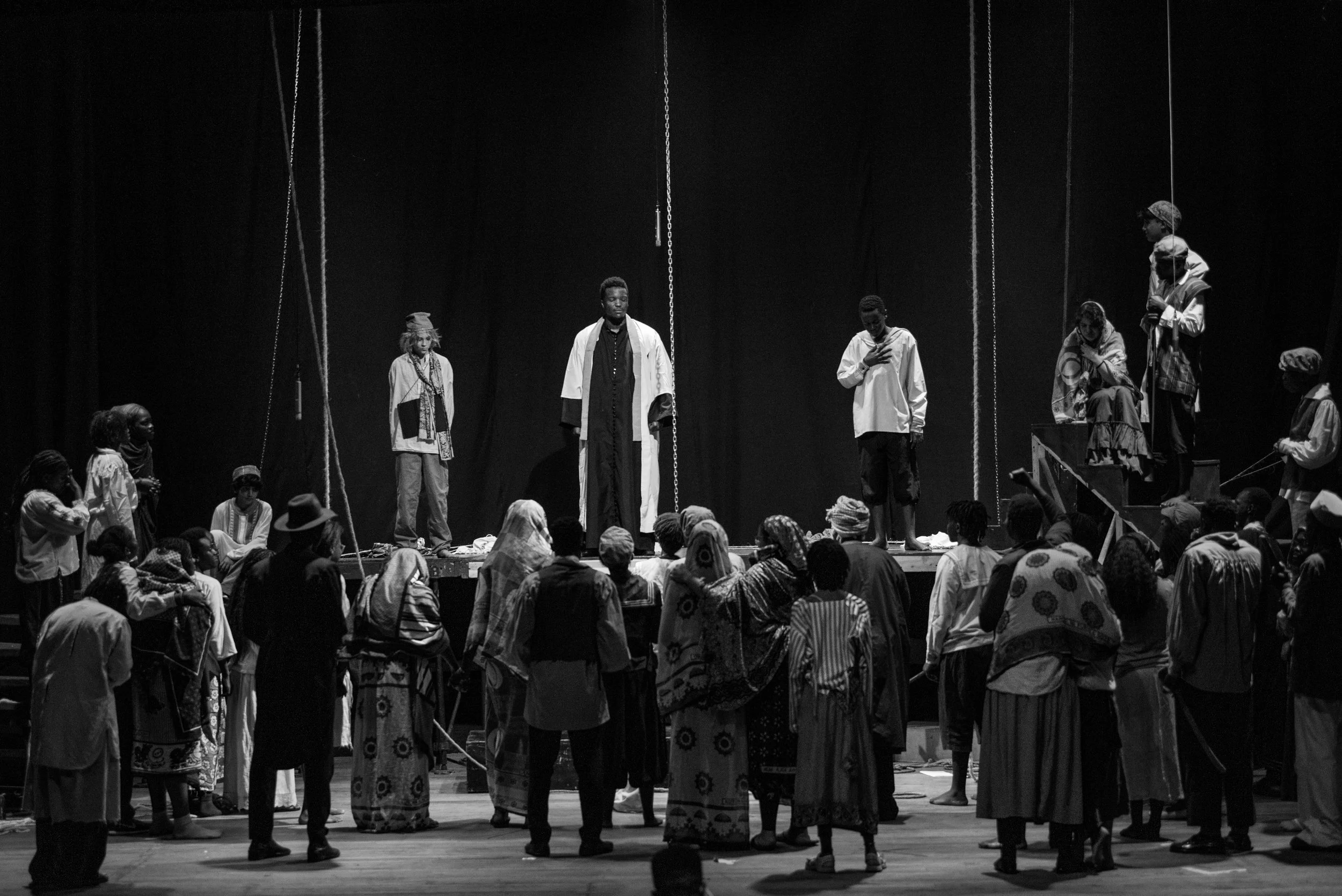 Black and white photo of a theatrical performance with multiple actors on stage dressed in costumes representing different characters. The audience is watching the scene, with some people on the stage and some standing below.