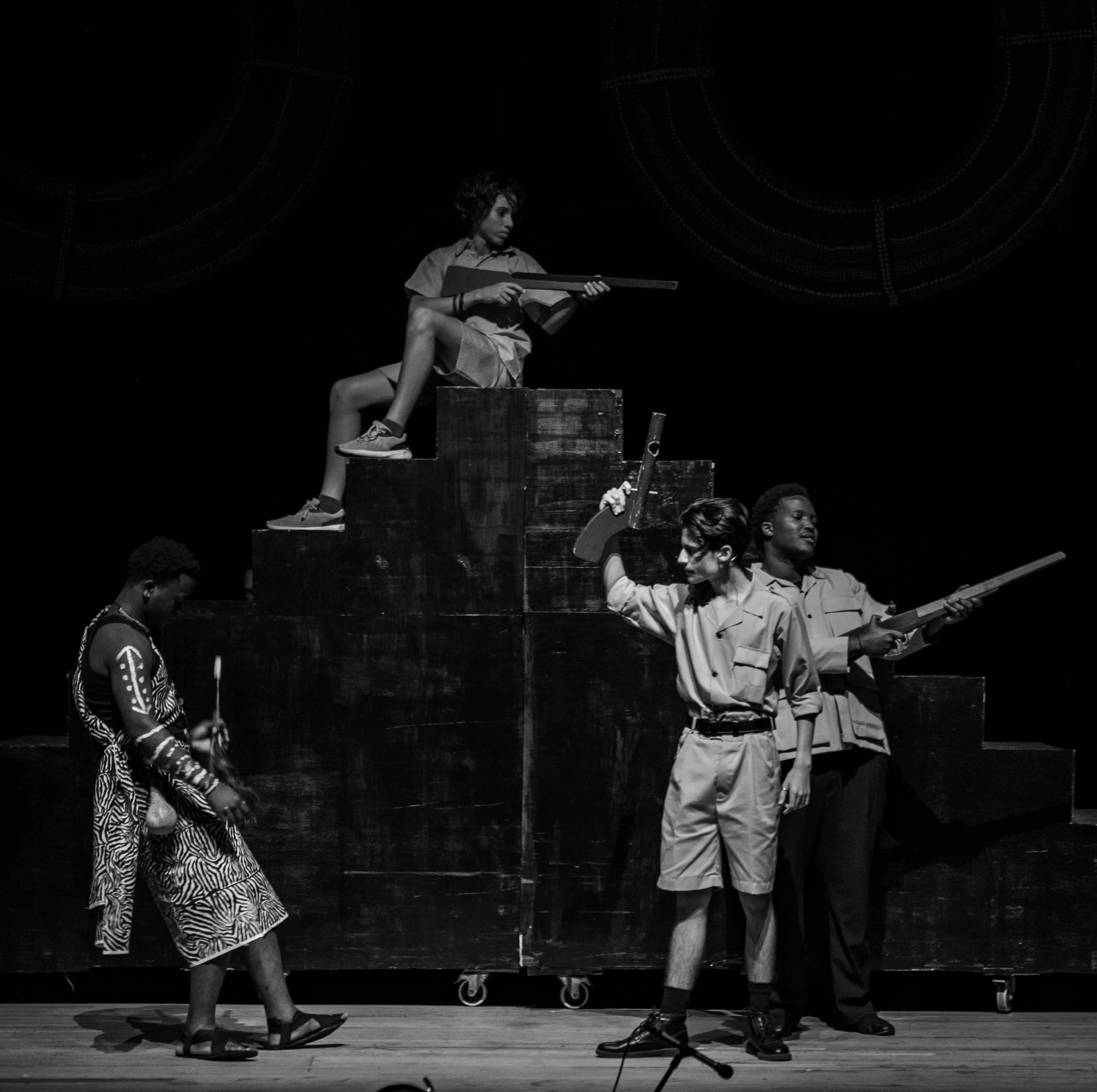 Black and white photo of four children on stage, some holding toy guns, in a theatrical performance or play.