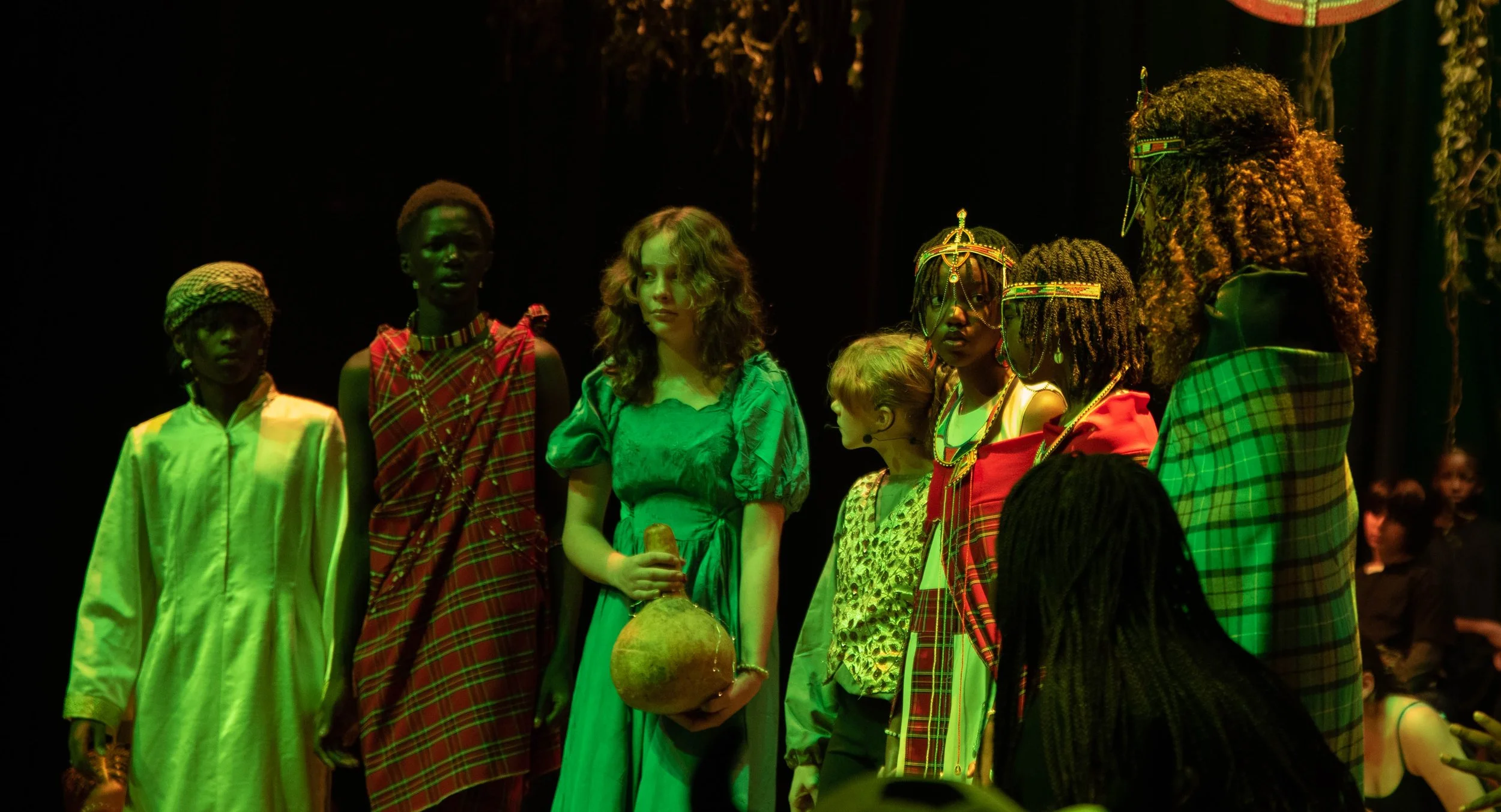 A group of diverse people standing on a stage with dark background, some dressed in traditional African clothing, and a woman holding a gourd in the center.