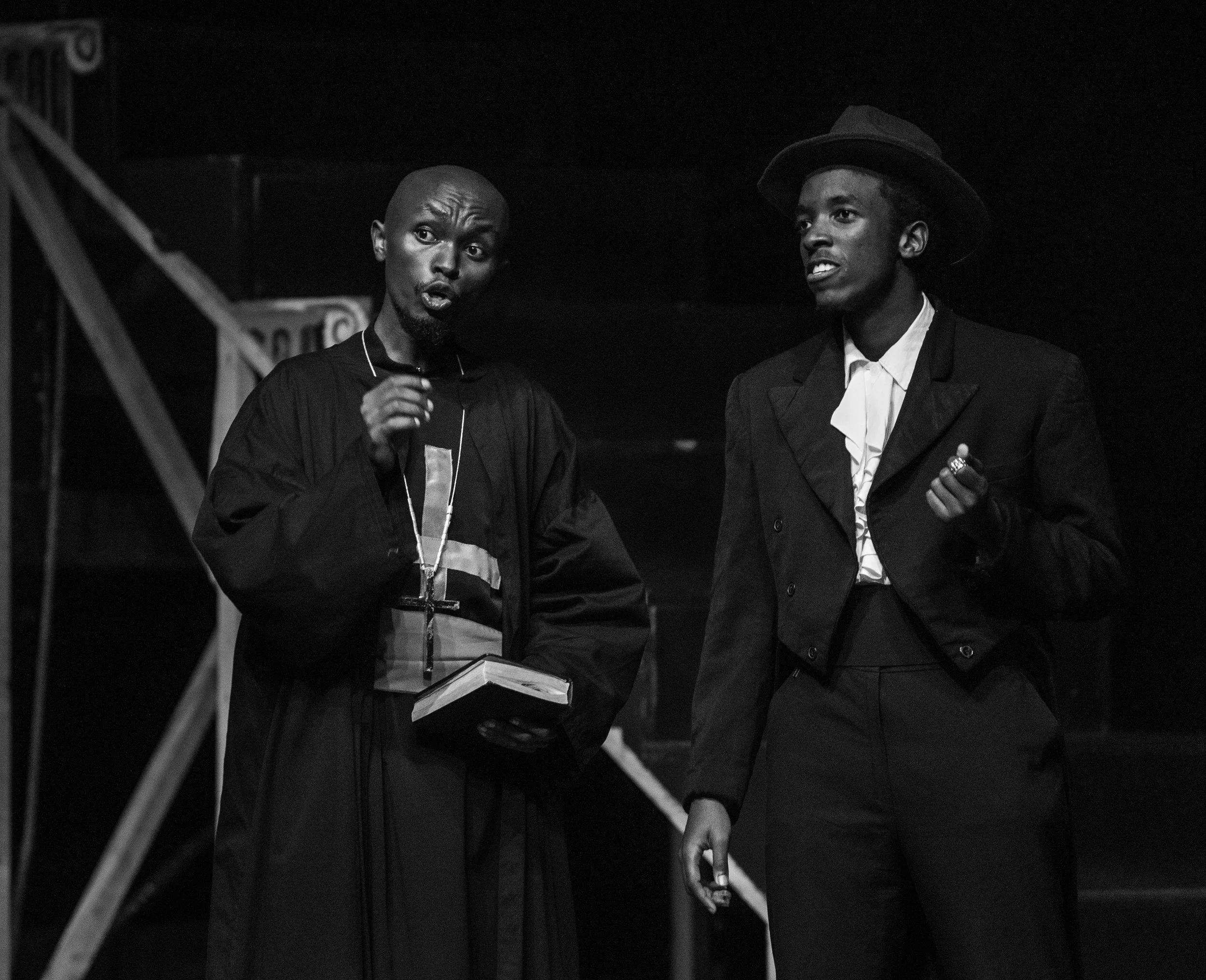 Two men, one in a robe holding a book, and the other in a suit and hat, are engaged in a conversation on a stage.