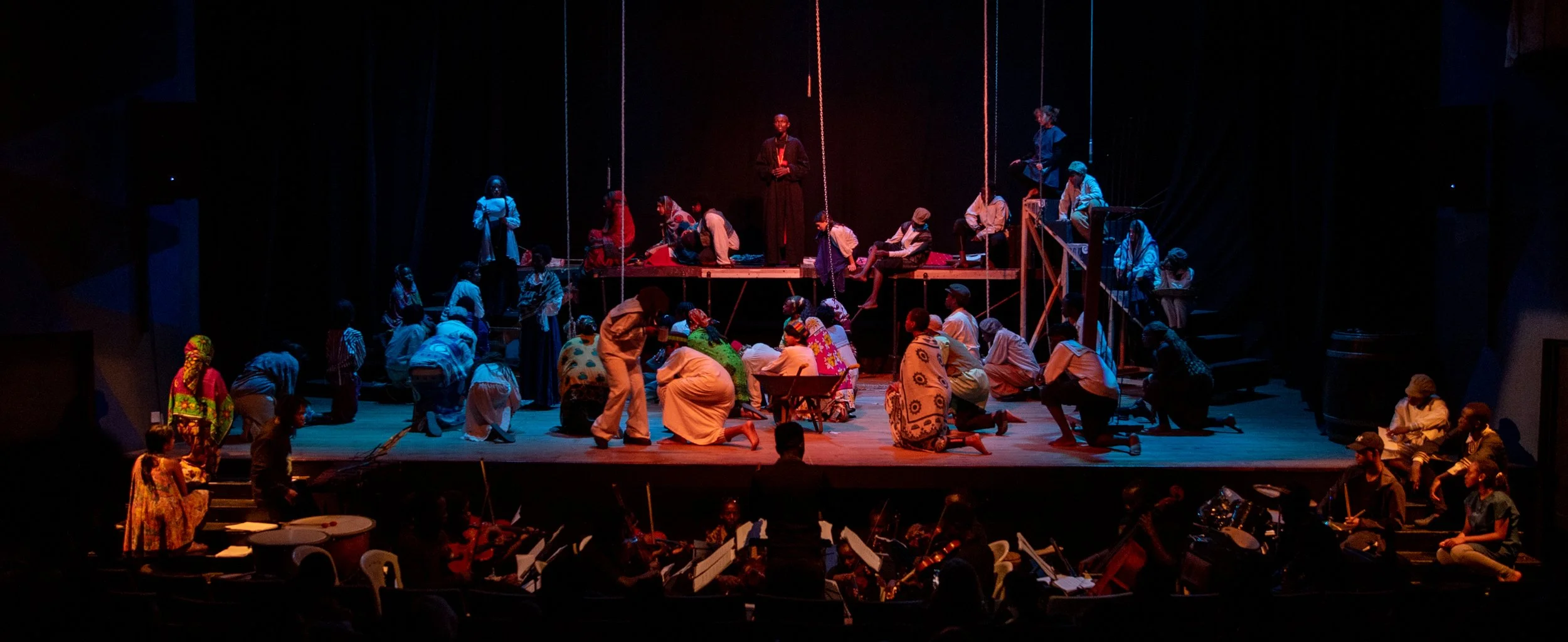 Theater stage with actors in traditional costumes performing, musicians playing instruments in front, and a conductor leading an orchestra.
