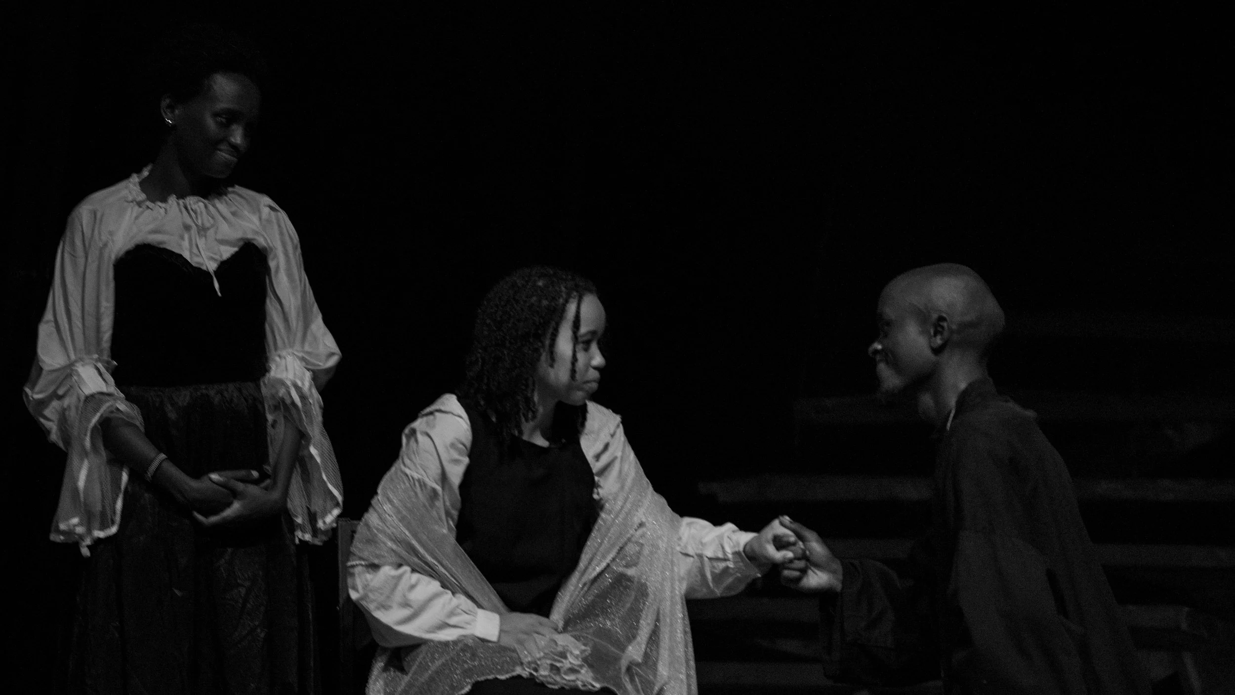 Three women in a dark setting. One woman is standing while the other two are sitting and holding hands, engaging in conversation or a gesture.