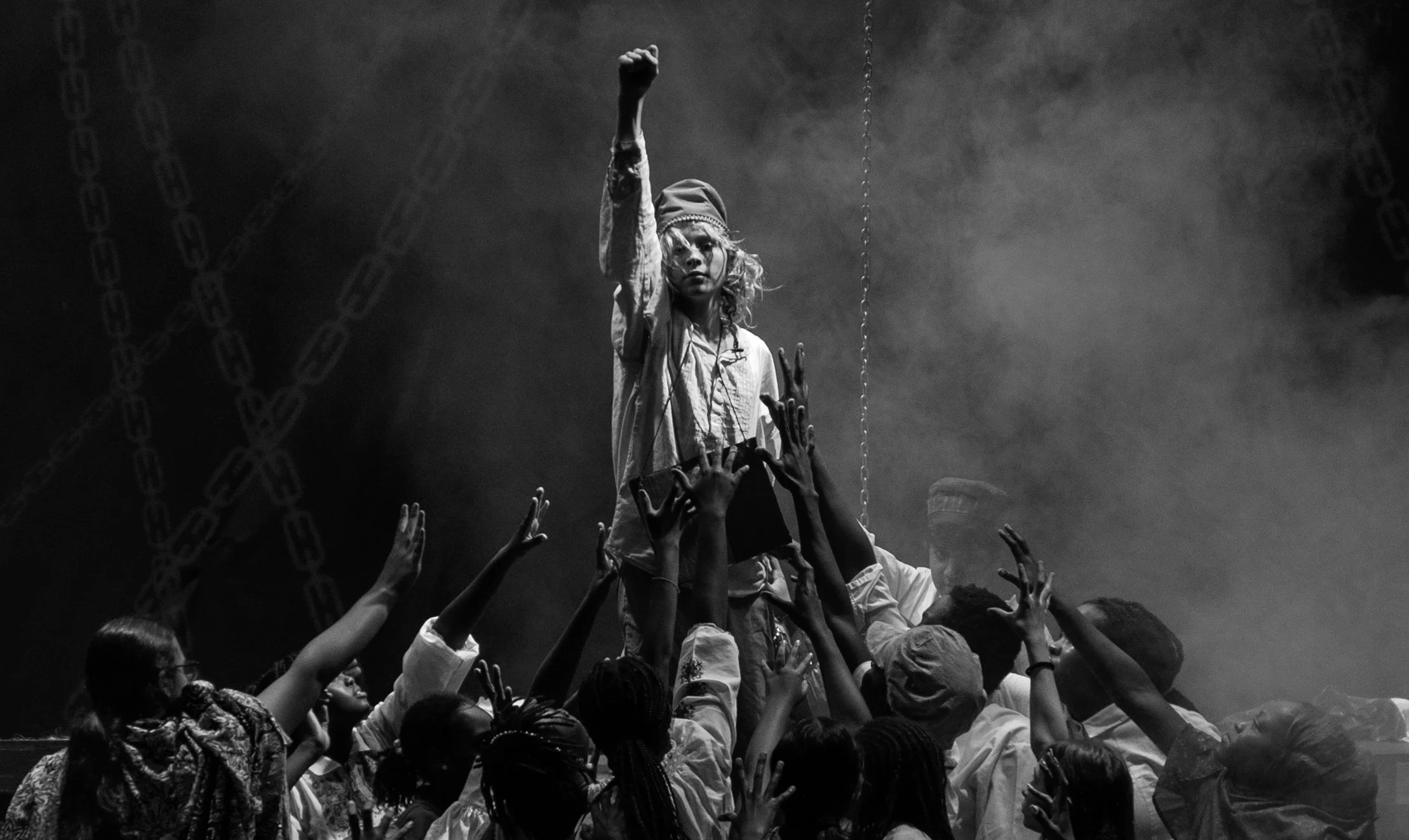 A black-and-white photo of a woman standing on a platform amid a crowd, with her right arm raised and tights into a fist. Several people are reaching toward her with their hands. The scene appears intense and dramatic.