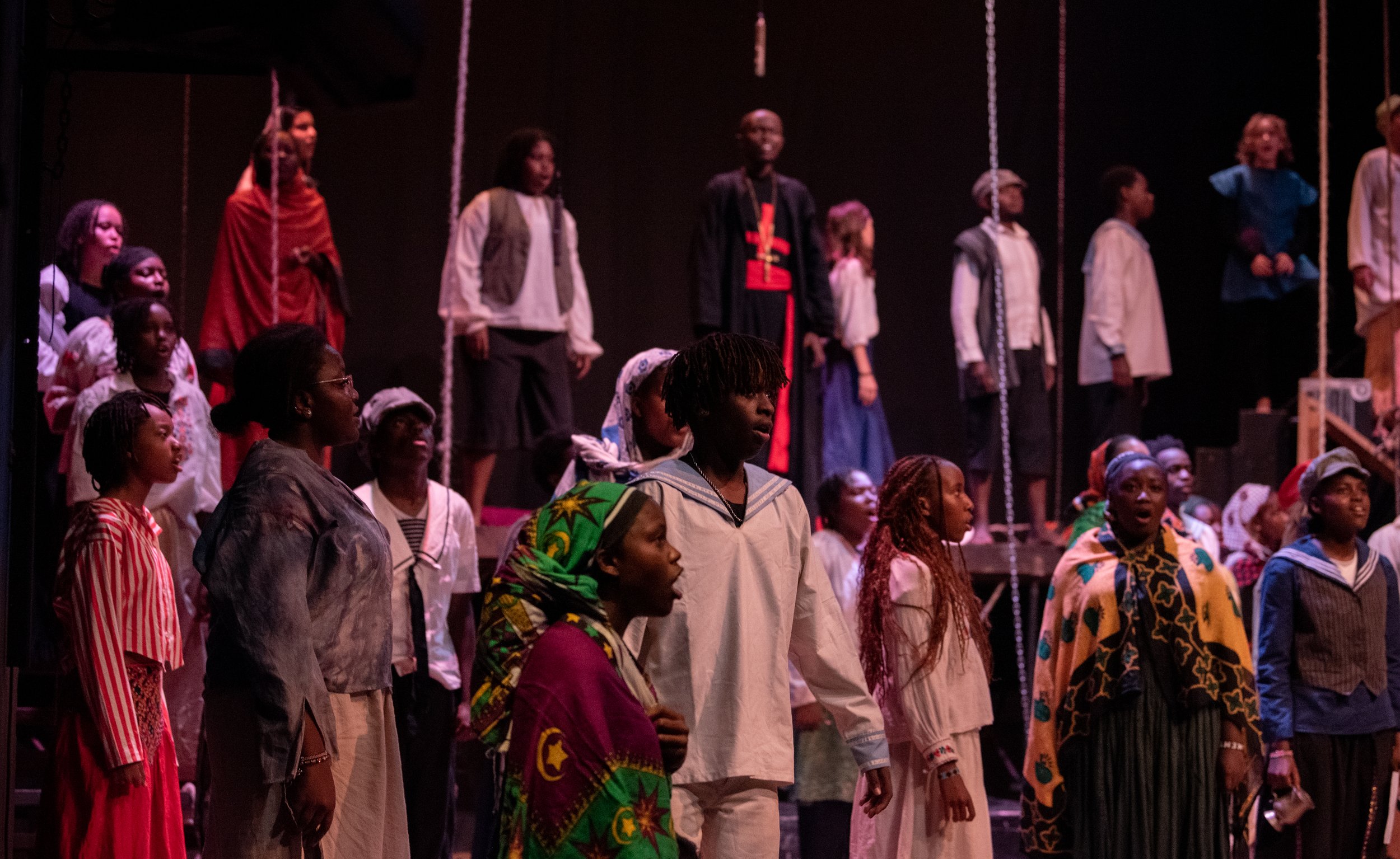 A diverse group of children and teenagers performing on stage, some standing and singing with serious expressions, dressed in a mix of casual and traditional clothing.