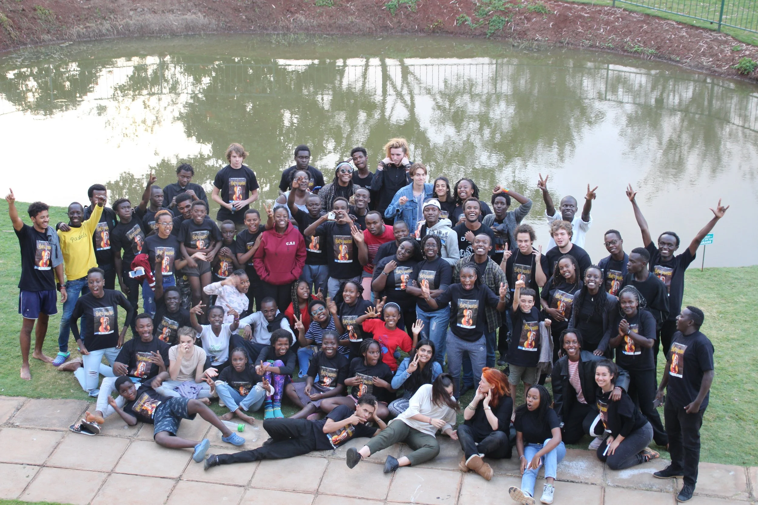 A large group of diverse people, including children, teenagers, and adults, gathered outdoors near a pond, posing for a group photo with many smiling and making peace signs.