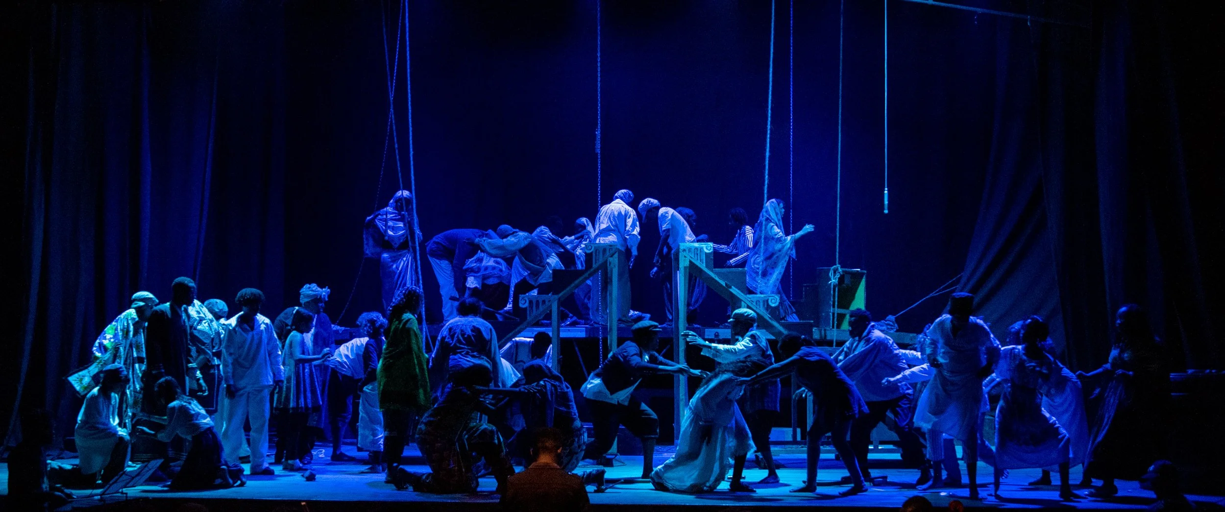 The image depicts a theatrical stage performance with many actors dressed in traditional South Asian attire, engaging in a dramatic scene under blue lighting. The scene includes multiple levels and actors reaching out to each other, with a dark curta