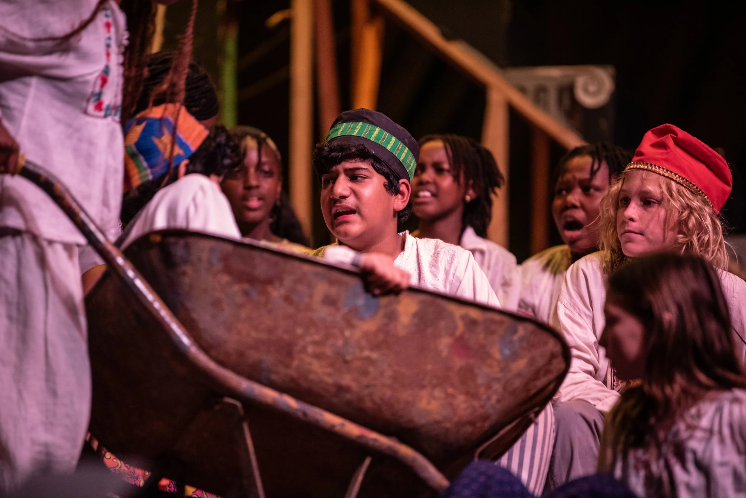 Children dressed in historical or traditional clothing, gathered around a large wheelbarrow or similar object, with some showing expressions of concern or concentration, participating in a group activity or performance in a dimly lit setting.