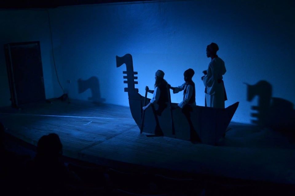 Children performing a play on stage with a boat set, lit with blue lighting, silhouette of children in costumes, some in traditional clothing, in a boat prop.