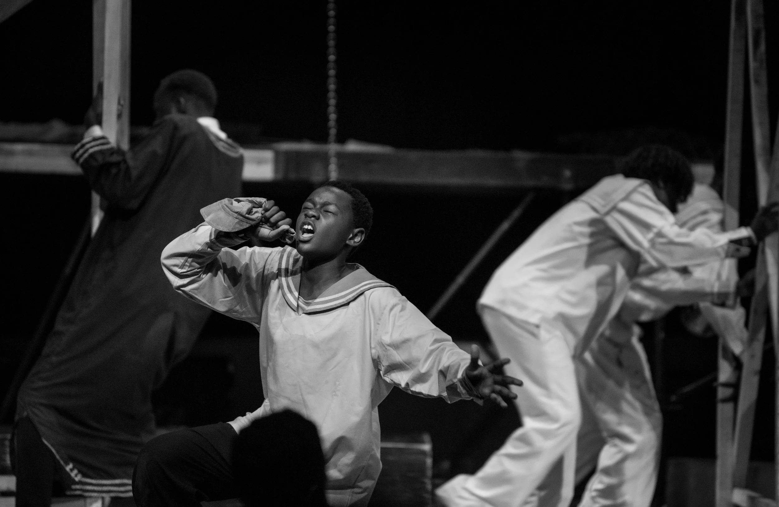 A boy singing passionately on stage, with two other people focused on their phones, in a black and white photo.