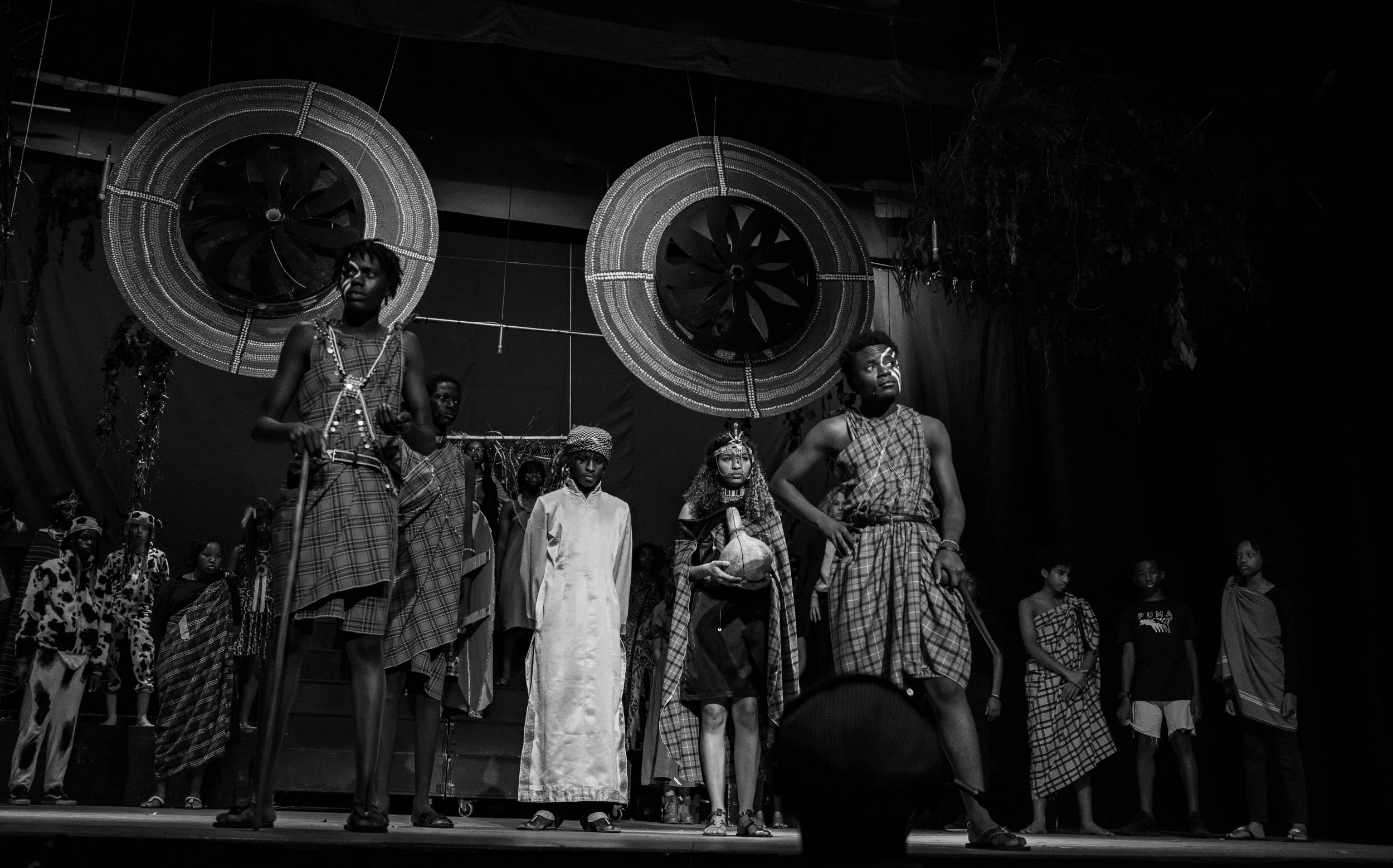 Group of people dressed in traditional African clothing on a stage with large circular decorations in the background.