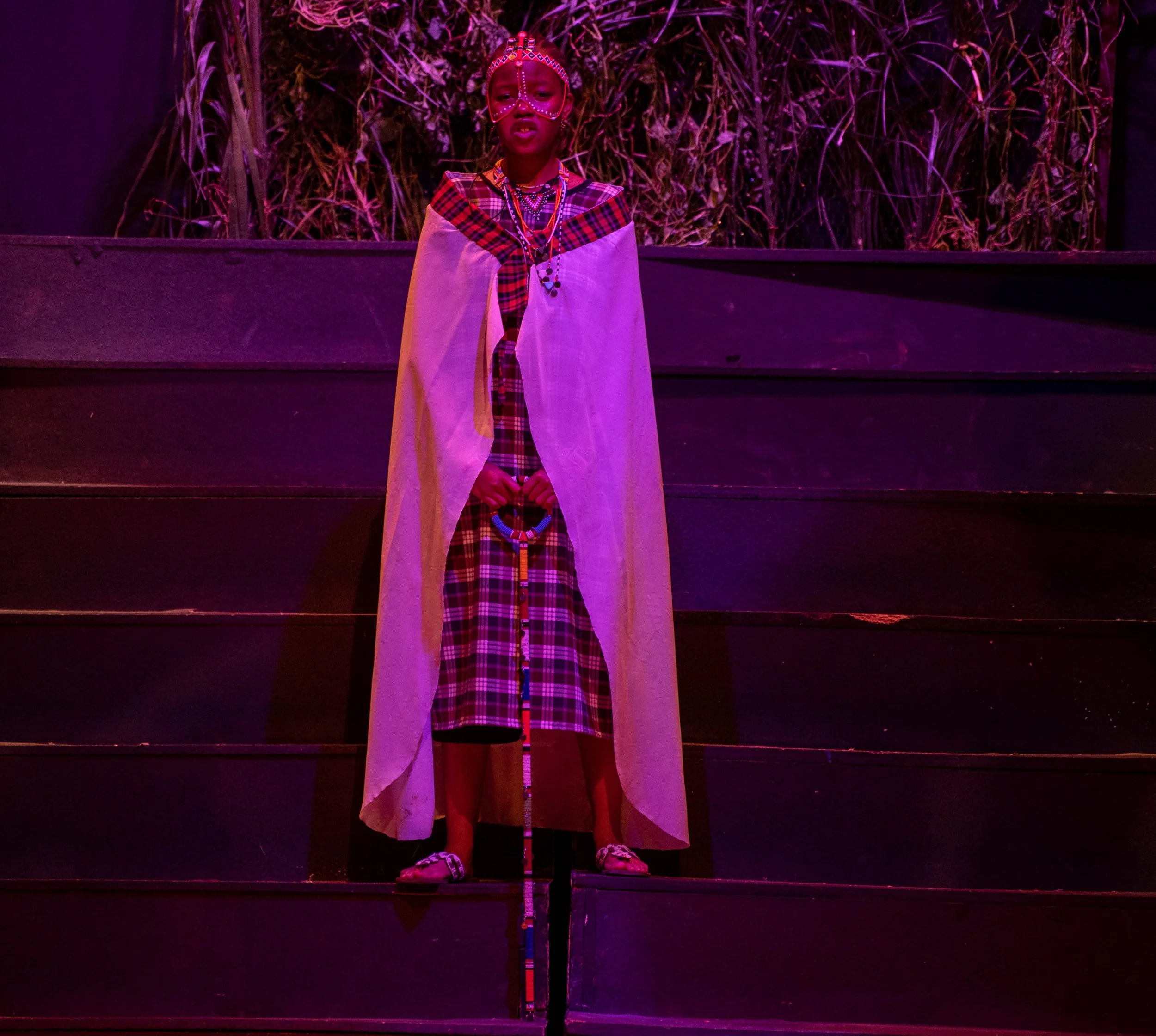 A young girl dressed in traditional African attire with face paint and jewelry, standing on stage with purple lighting and a background of plants.