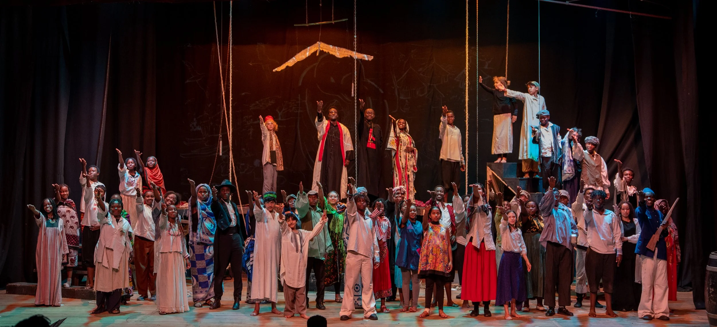 A large group of performers on stage during a theatrical production, with children and adults dressed in traditional and historical costumes, some standing and some sitting, and a dark background with theatrical lighting.