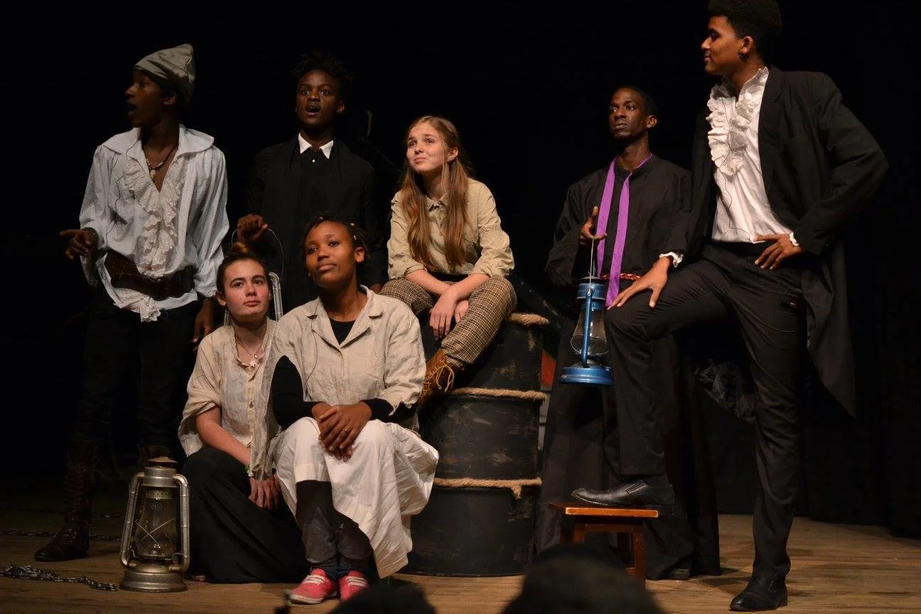 Group of seven young actors on stage in a theatrical play, dressed in period costumes, with some sitting and others standing, against a dark background.