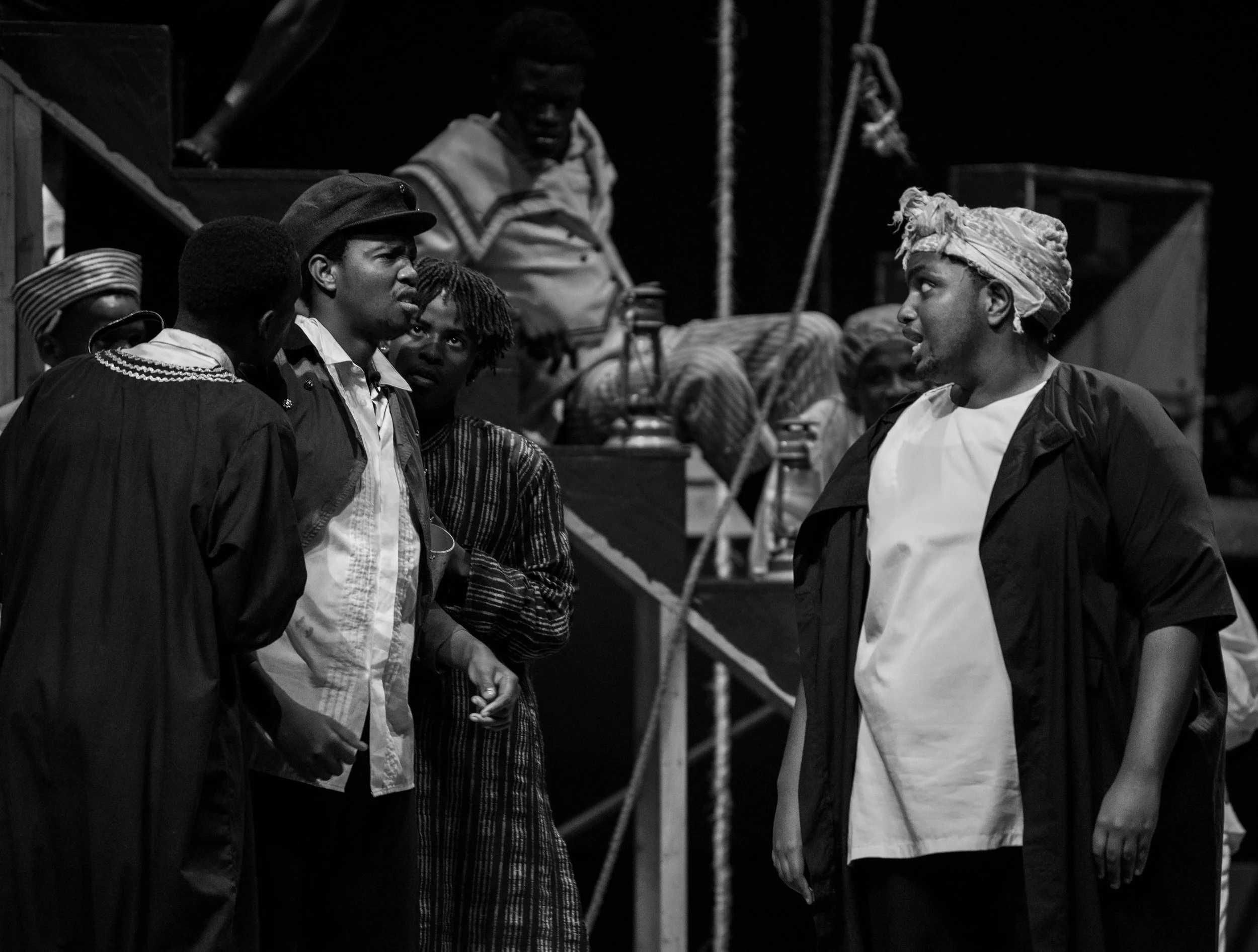 A black and white scene from a theatrical performance depicting several actors engaged in an intense conversation on stage, with some actors in costumes and headwear.