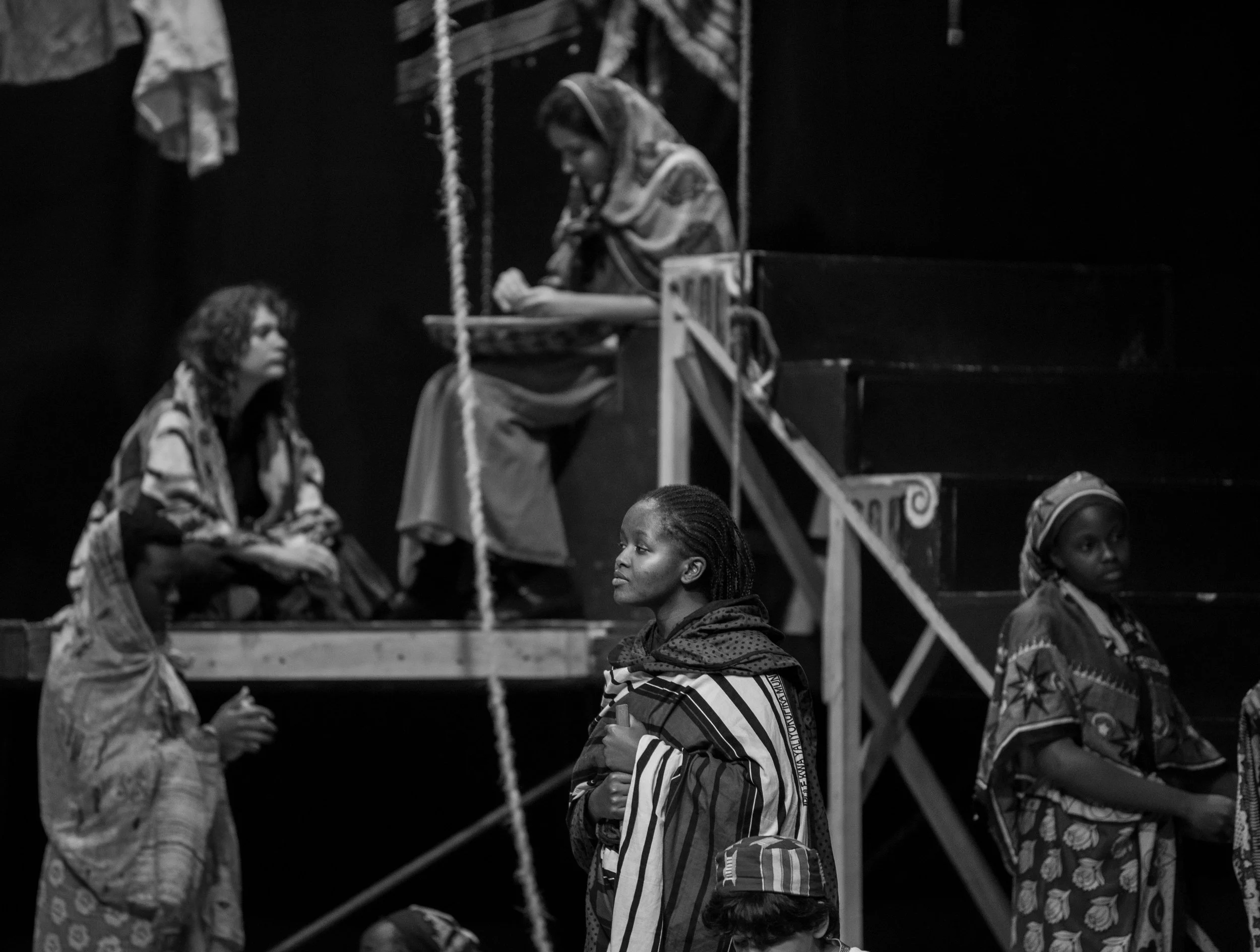 Black and white photo of women on stage, some standing and some sitting, dressed in patterned clothing with headscarves, against a dark background.