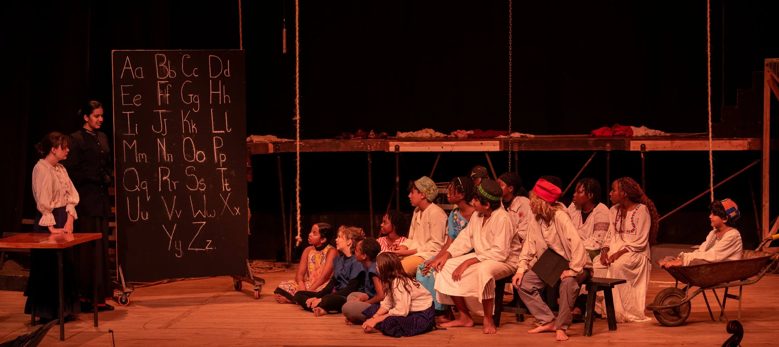 Children sitting on a stage during a school play, with an adult standing next to a blackboard displaying the alphabet, in a setting resembling a classroom or educational environment.